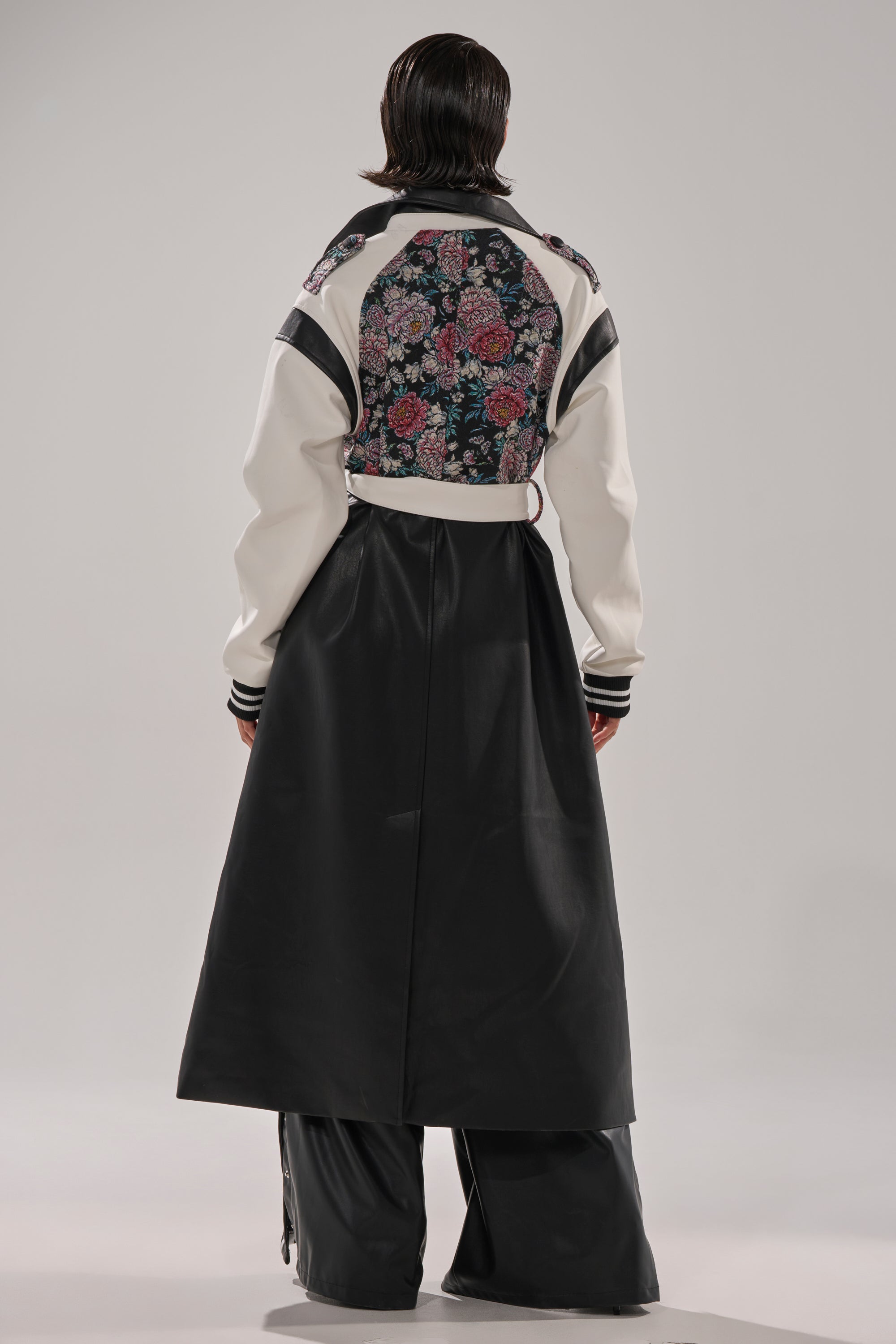 A person with slicked-back dark hair stands facing away in the END GAME MIXED MEDIA TRENCH, a long black and white coat featuring a floral pattern on the upper back, paired with wide-leg black pants against a light background.