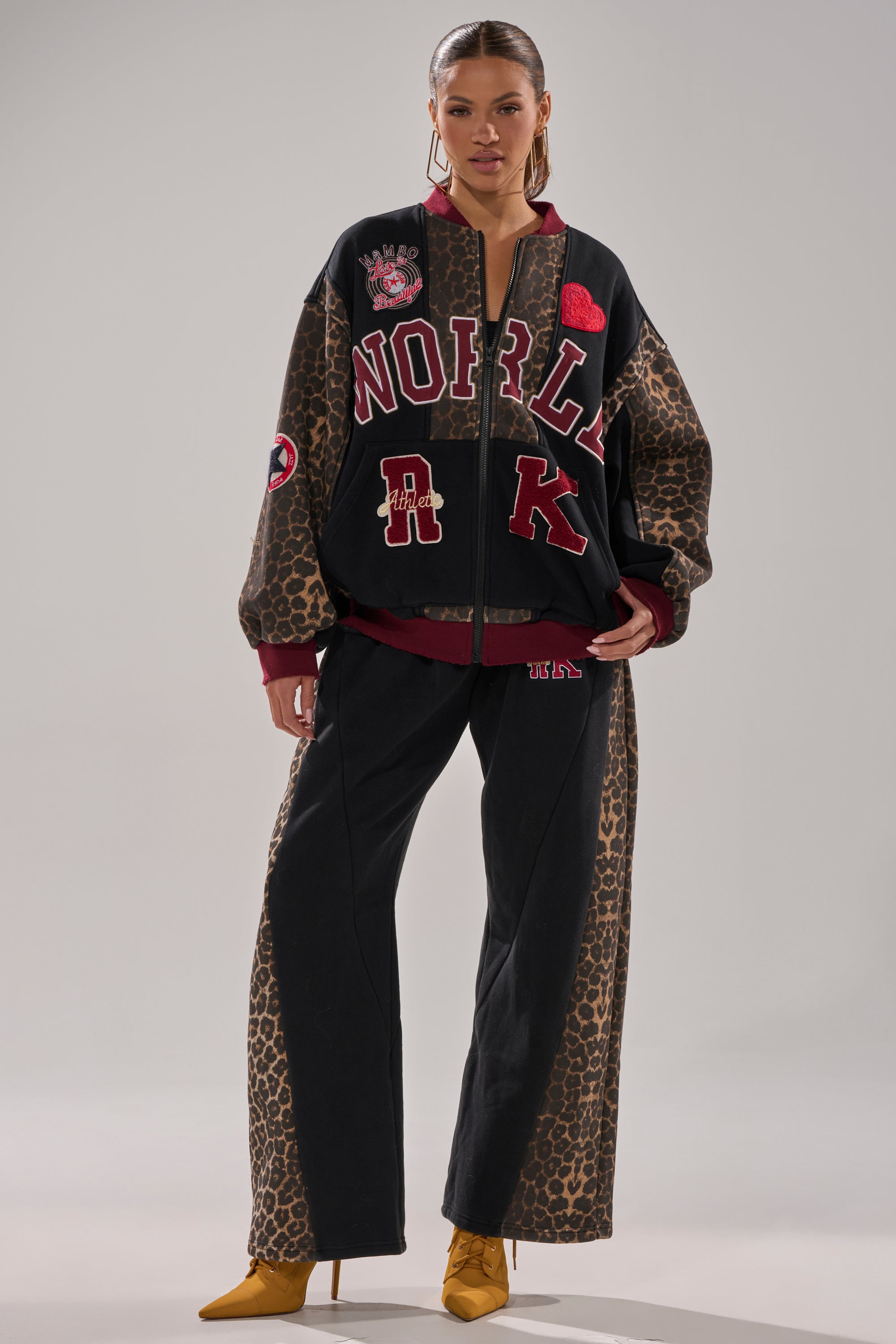 A woman faces forward wearing the WORLD HOLD ON JOGGER set—loose leopard-print and black jacket with red and white patches, matching joggers, and yellow heeled boots against a plain background.
