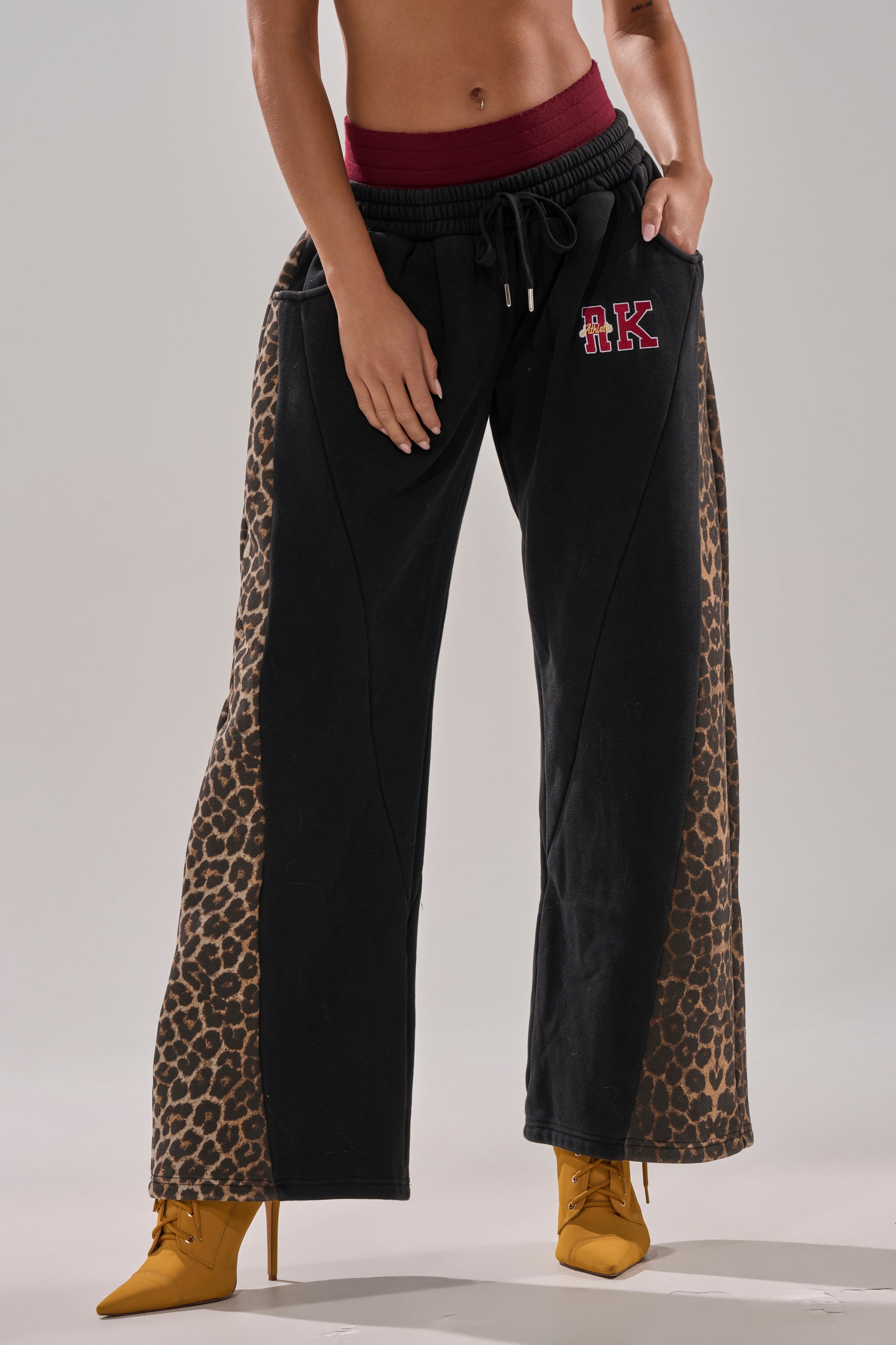 A person wears the WORLD HOLD ON JOGGER featuring black leopard print, a red waistband, and "RK" lettering on the left thigh, paired with yellow heeled boots. The upper body is cropped out of the image.
