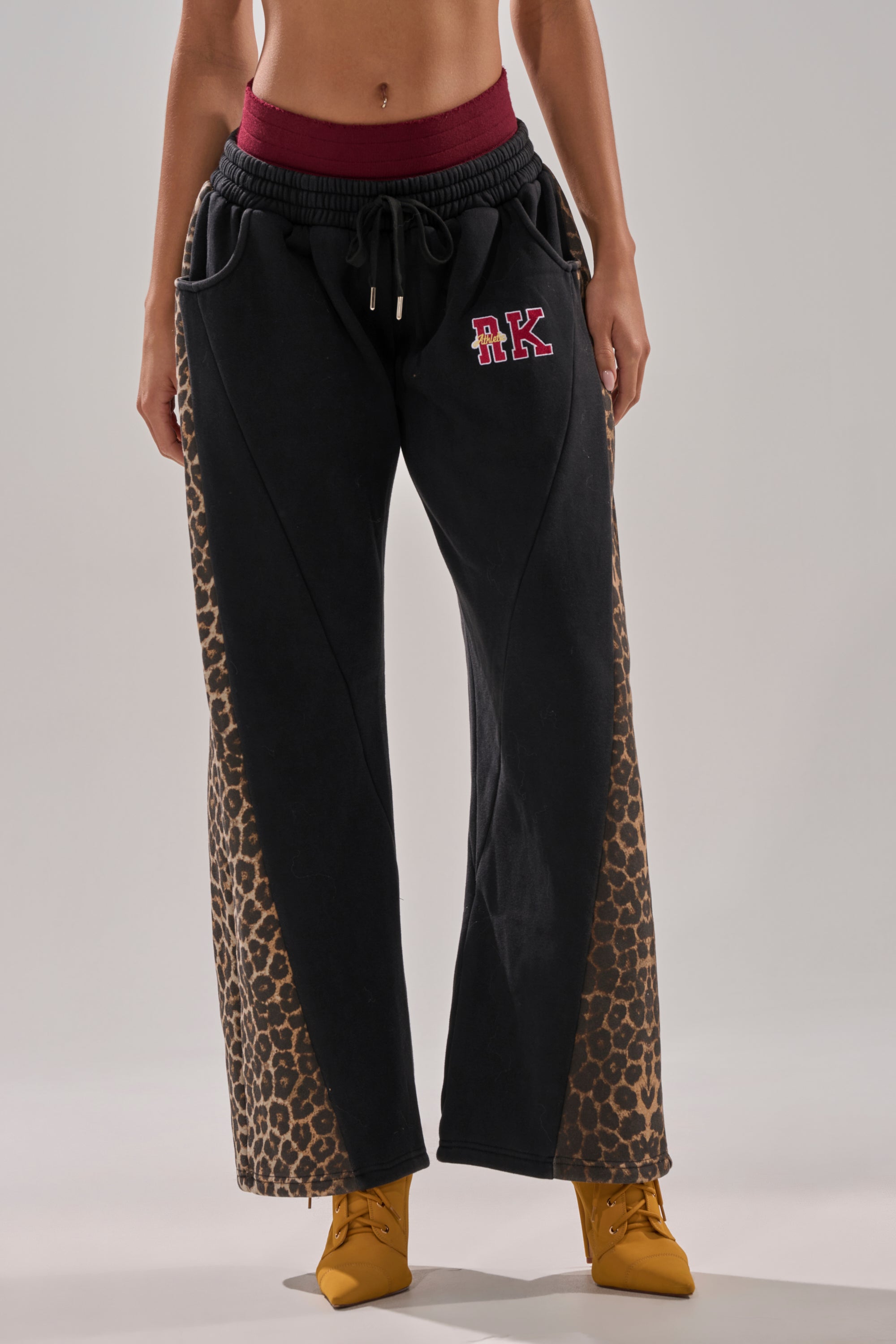 Wearing WORLD HOLD ON JOGGER sweatpants with relaxed fit, leopard print side panels, maroon waistband, embroidered “RK” logo, and yellow pointed-toe boots, a person stands against a plain background.
