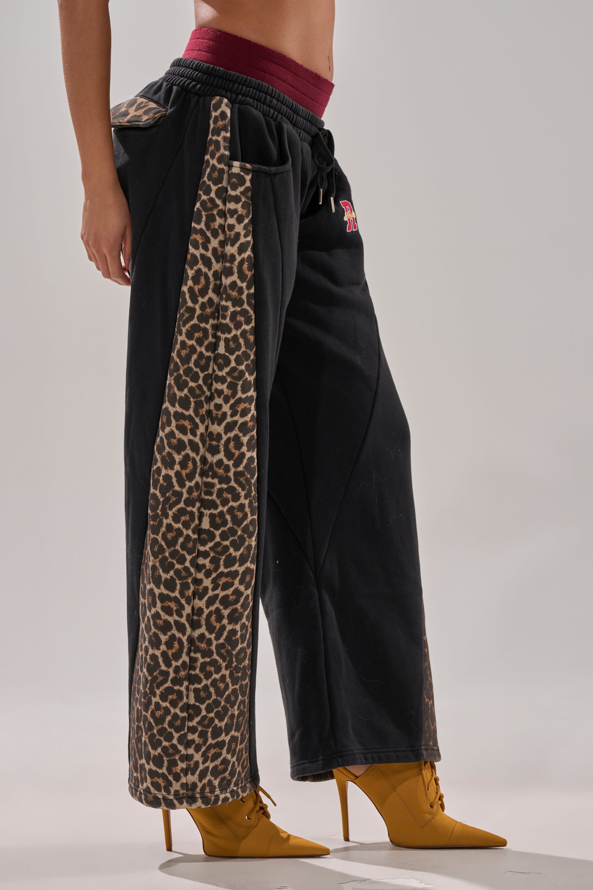 A person wears the WORLD HOLD ON JOGGER, featuring relaxed fit and leopard print side panels, paired with mustard yellow high-heeled ankle boots, posing against a light gray background.