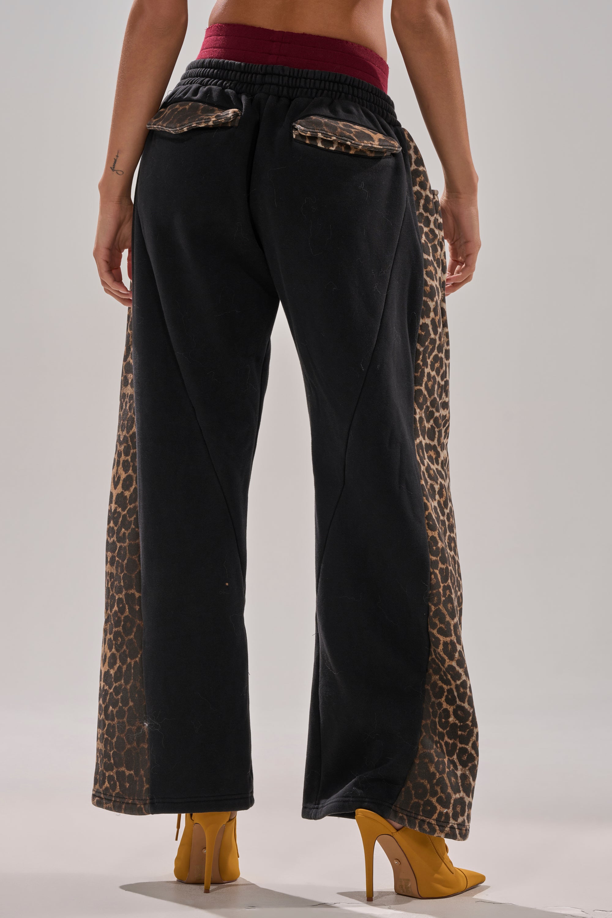 Someone wearing the WORLD HOLD ON JOGGER—black pants with leopard print side panels, back pockets, a burgundy waistband, and yellow heels—is photographed from behind.