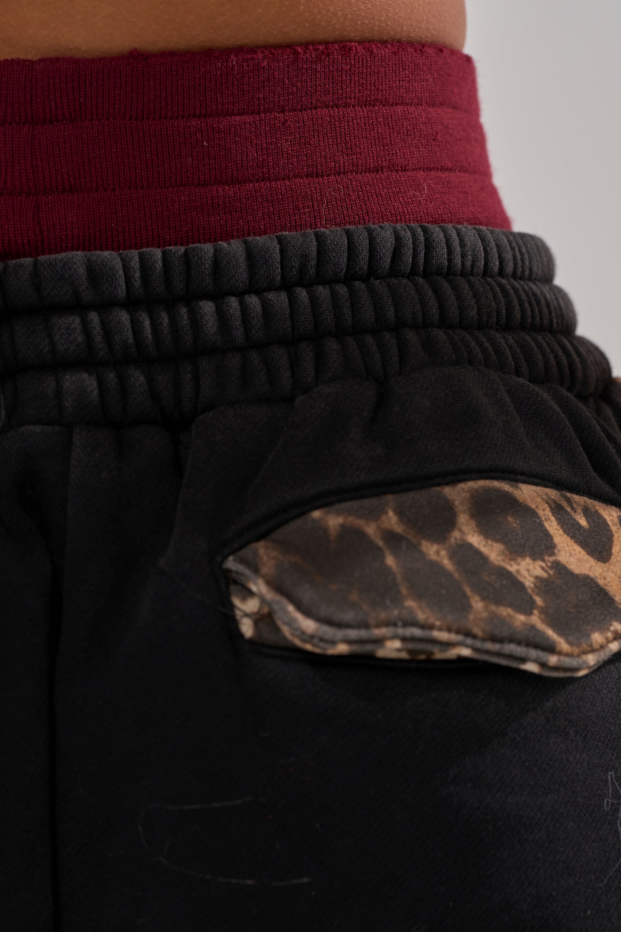 Close-up of a person wearing the WORLD HOLD ON JOGGER: black fleece sweatpants with an elastic waistband, a red waistband beneath, and a pocket with a brown leopard print flap.