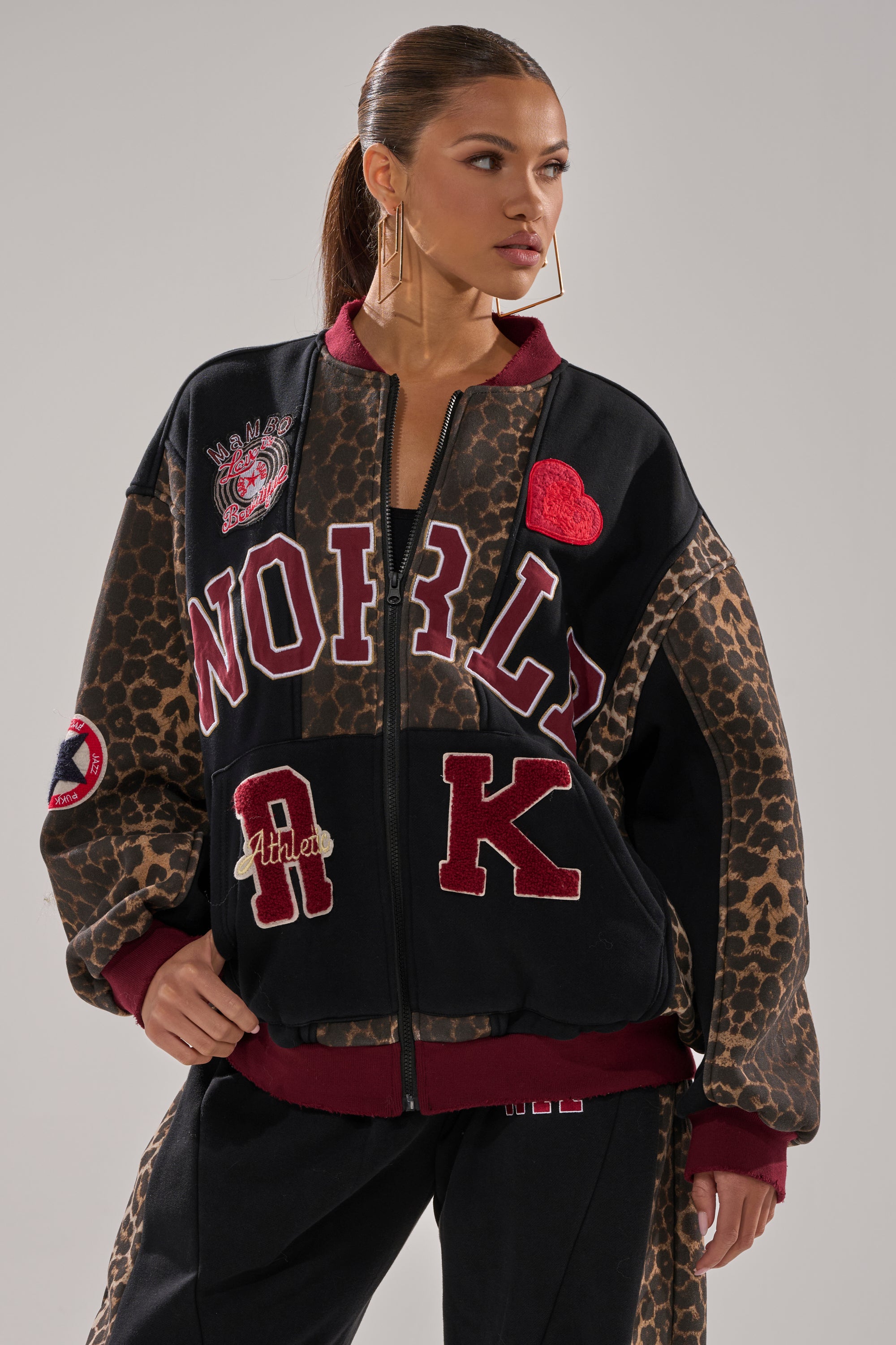 A woman wearing the WORLD HOLD ON ZIP UP SWEATSHIRT, featuring a leopard print and black varsity style with maroon patches, plus matching pants and large hoop earrings, stands with one hand in her pocket looking to the side.