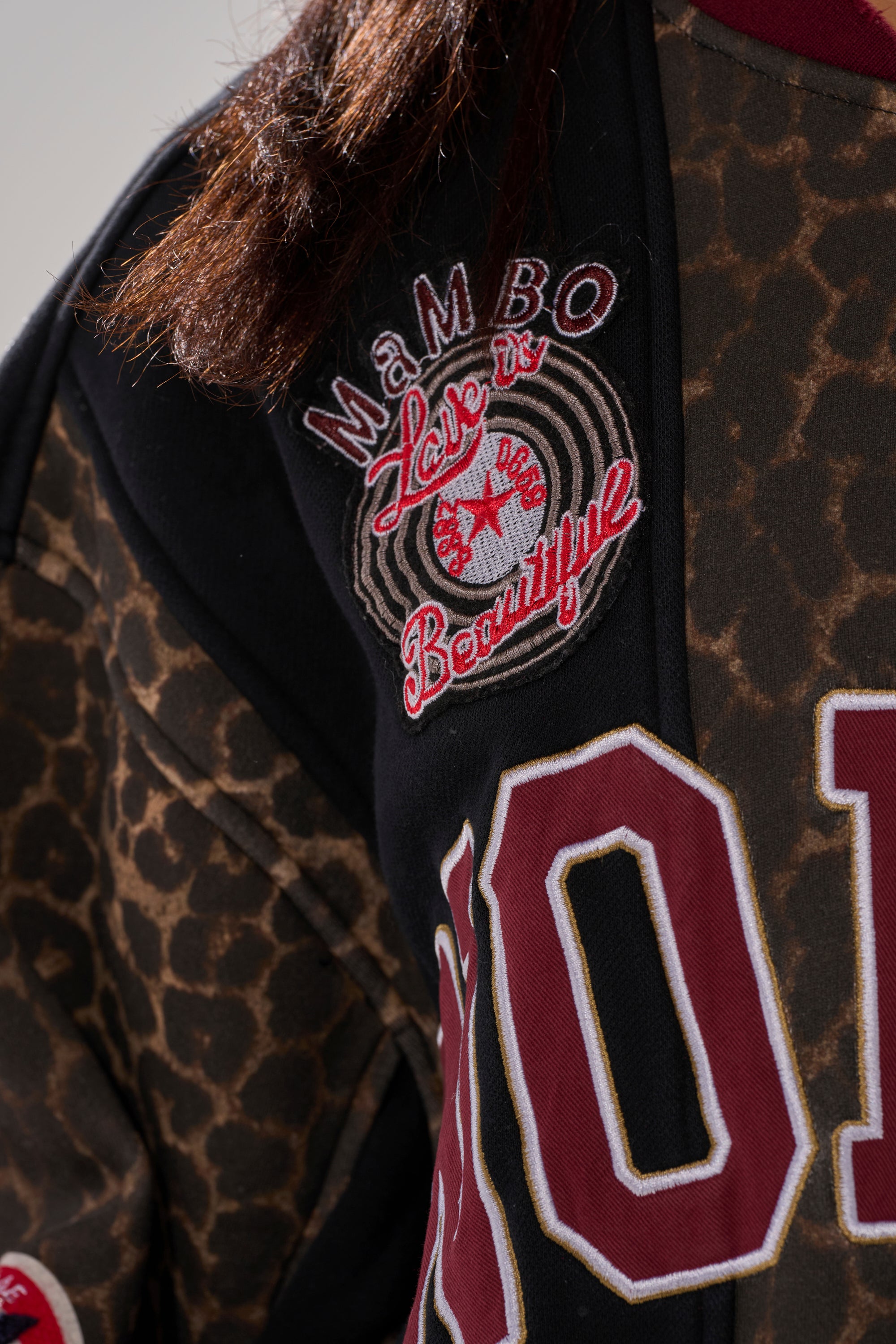 A close-up of a shoulder wearing the WORLD HOLD ON ZIP UP SWEATSHIRT, featuring bold leopard print, varsity patches reading "MAMBO," "Love," and "Beautiful," plus part of a large maroon letter.