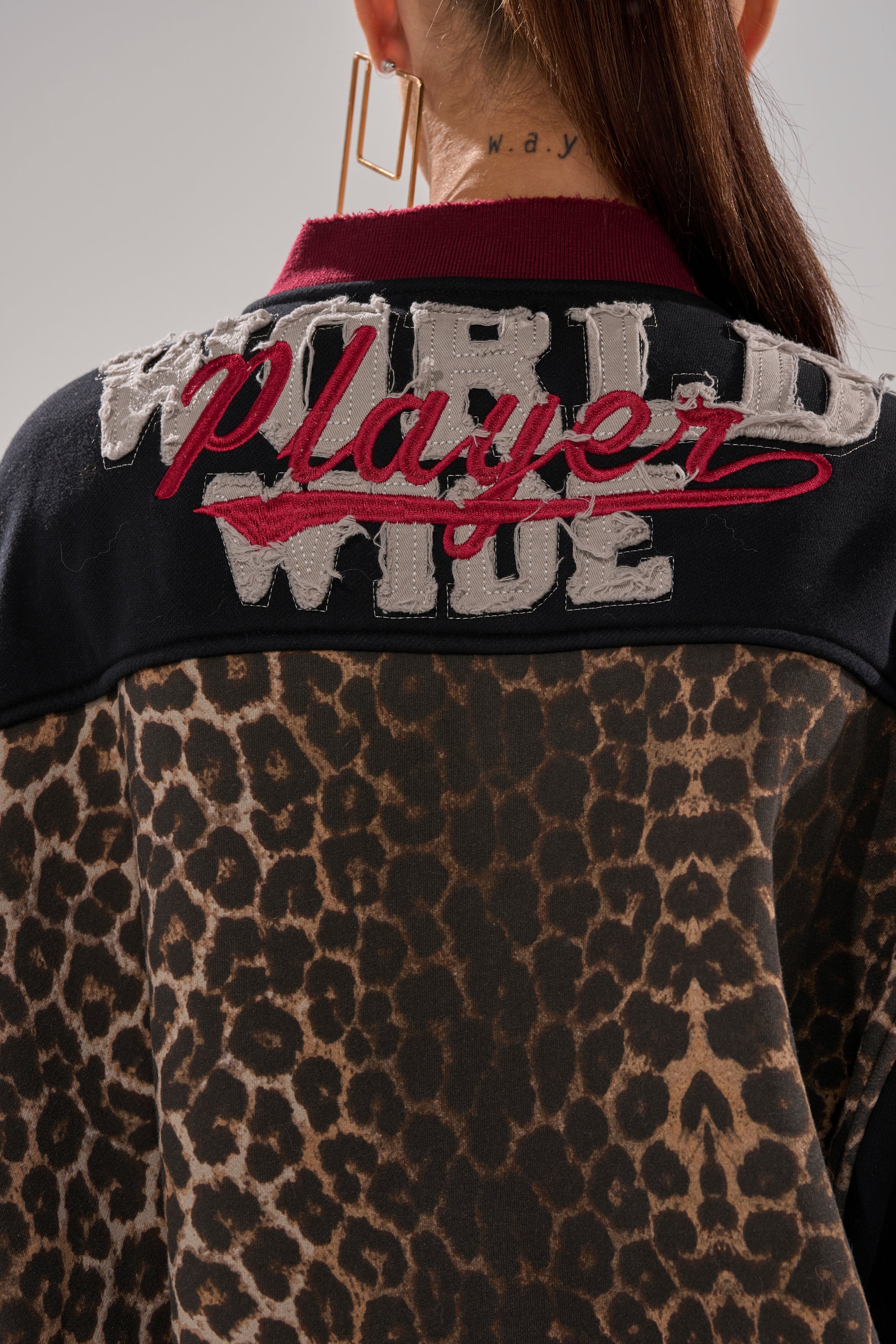 A person with an undercut tattoo reading “w.a.y.” wears the WORLD HOLD ON ZIP UP SWEATSHIRT, featuring a leopard print and bold embroidered letters on the back. Large gold earrings complete their look.