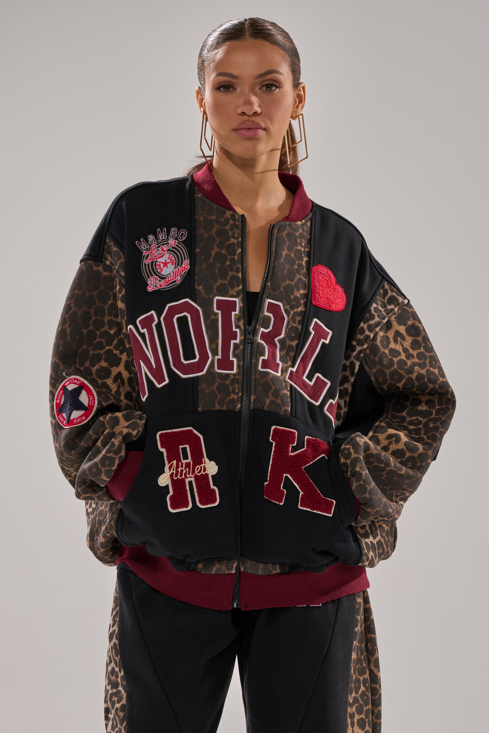 A woman stands confidently in the WORLD HOLD ON ZIP UP SWEATSHIRT featuring a loose leopard print with bold patches—“NORL,” “RK,” a red heart, and a sports emblem. She wears hoop earrings and has her hair pulled back against a plain gray background.