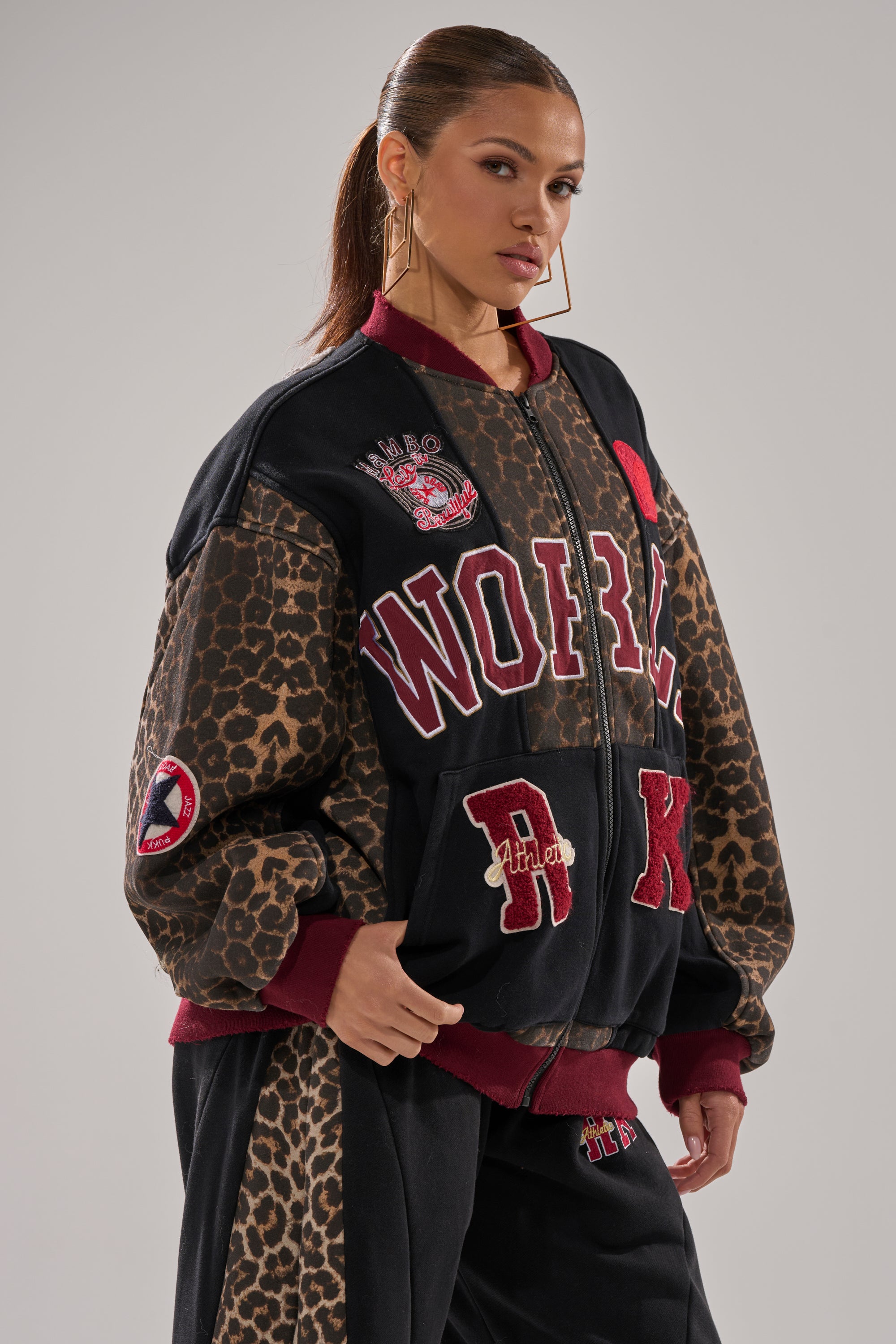 A woman with a high ponytail models the WORLD HOLD ON ZIP UP SWEATSHIRT, featuring black varsity style, bold leopard print panels, red and white lettering, patches, and large gold hoop earrings against a plain background.