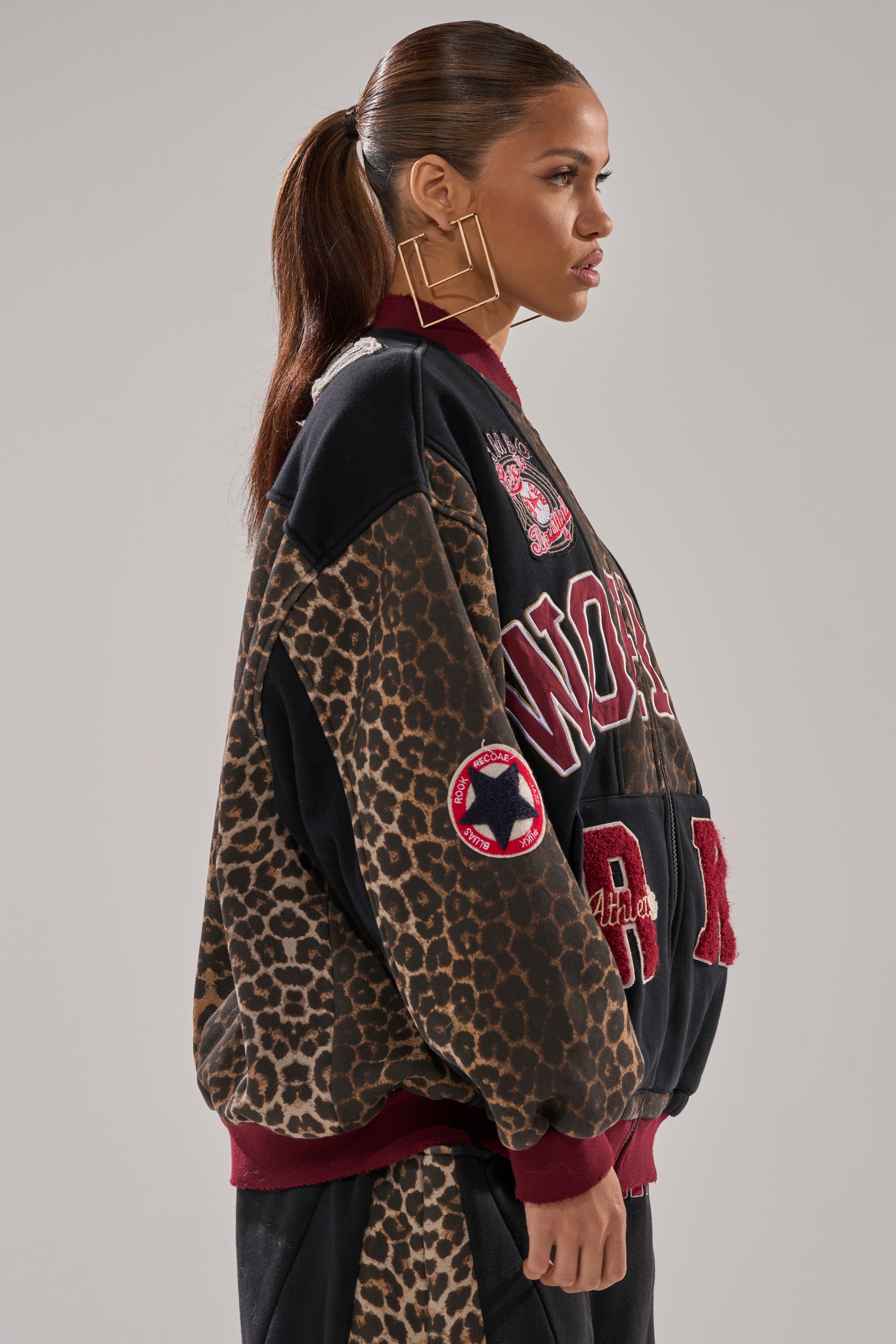 A woman with a sleek ponytail wears the WORLD HOLD ON ZIP UP SWEATSHIRT, featuring a leopard print, red accents, and bold patches, paired with large square earrings. She stands in profile against a plain background.