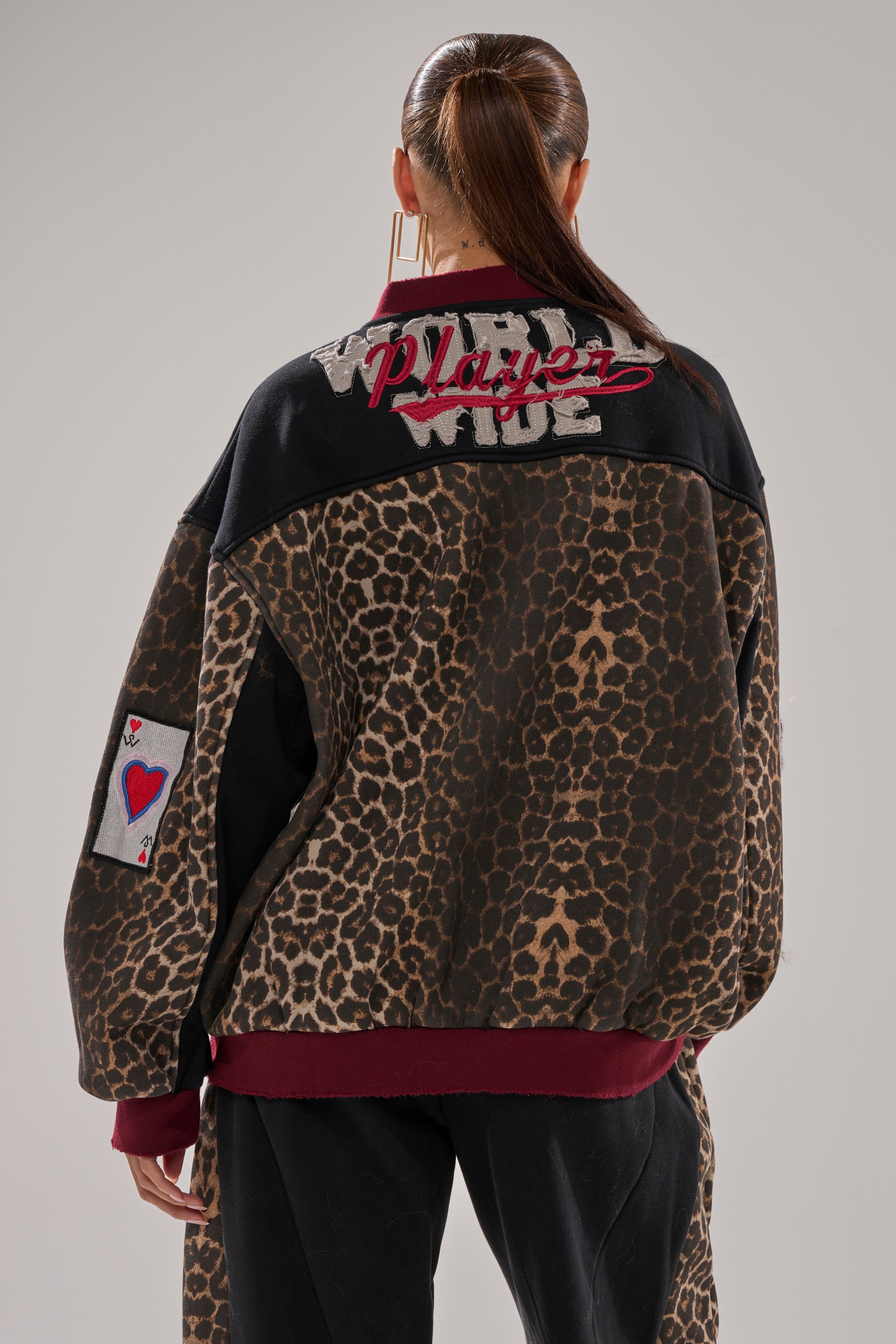 A person with a sleek ponytail wears the WORLD HOLD ON ZIP UP SWEATSHIRT, featuring varsity style leopard print, black sections, burgundy cuffs, and embroidered patches—“PLAYER” on the back and a heart playing card on the sleeve.