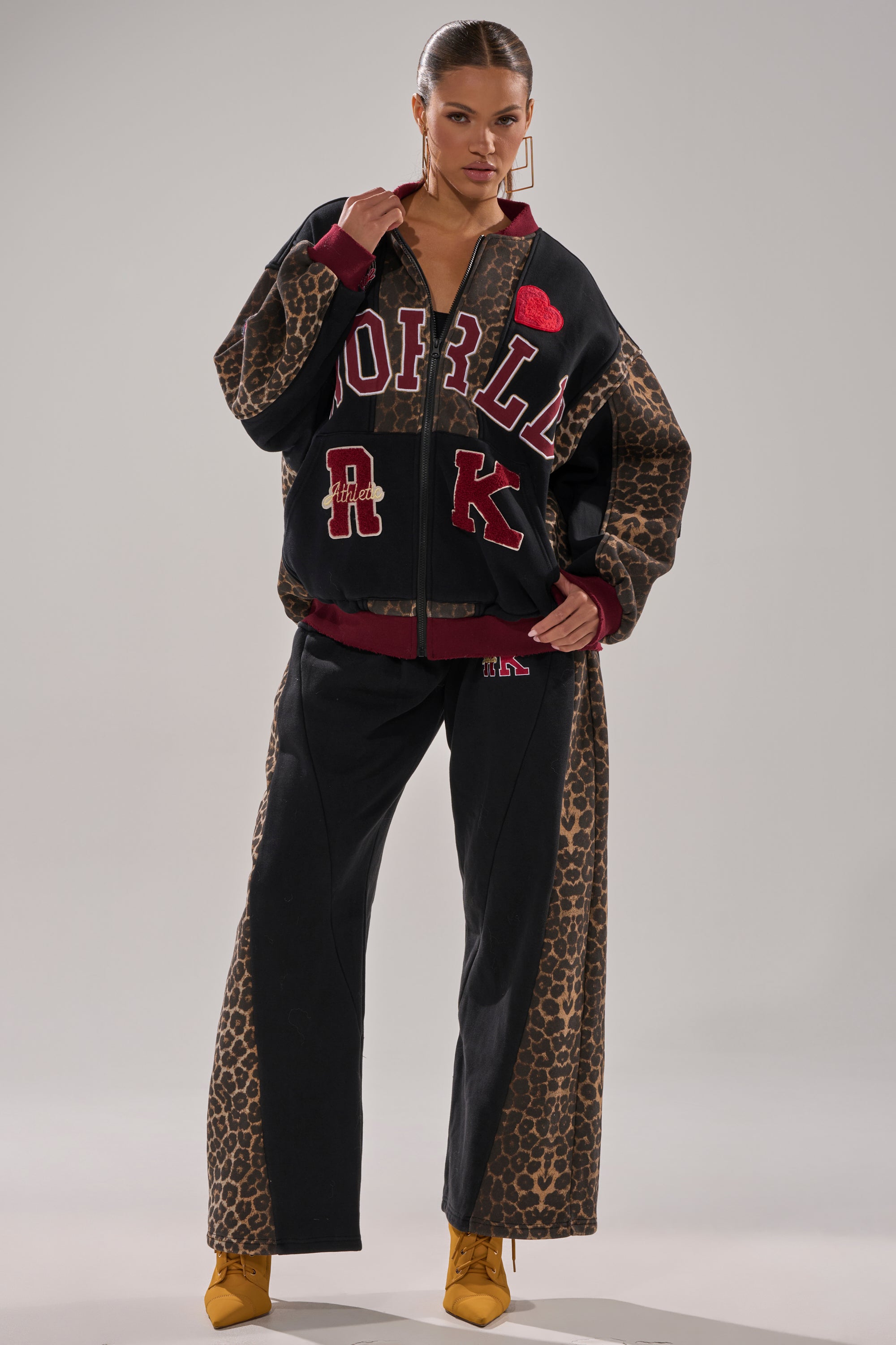 A woman stands with hands in pockets, wearing the WORLD HOLD ON ZIP UP SWEATSHIRT with black and leopard print varsity details, matching wide-leg pants, and yellow lace-up boots against a plain grey background.