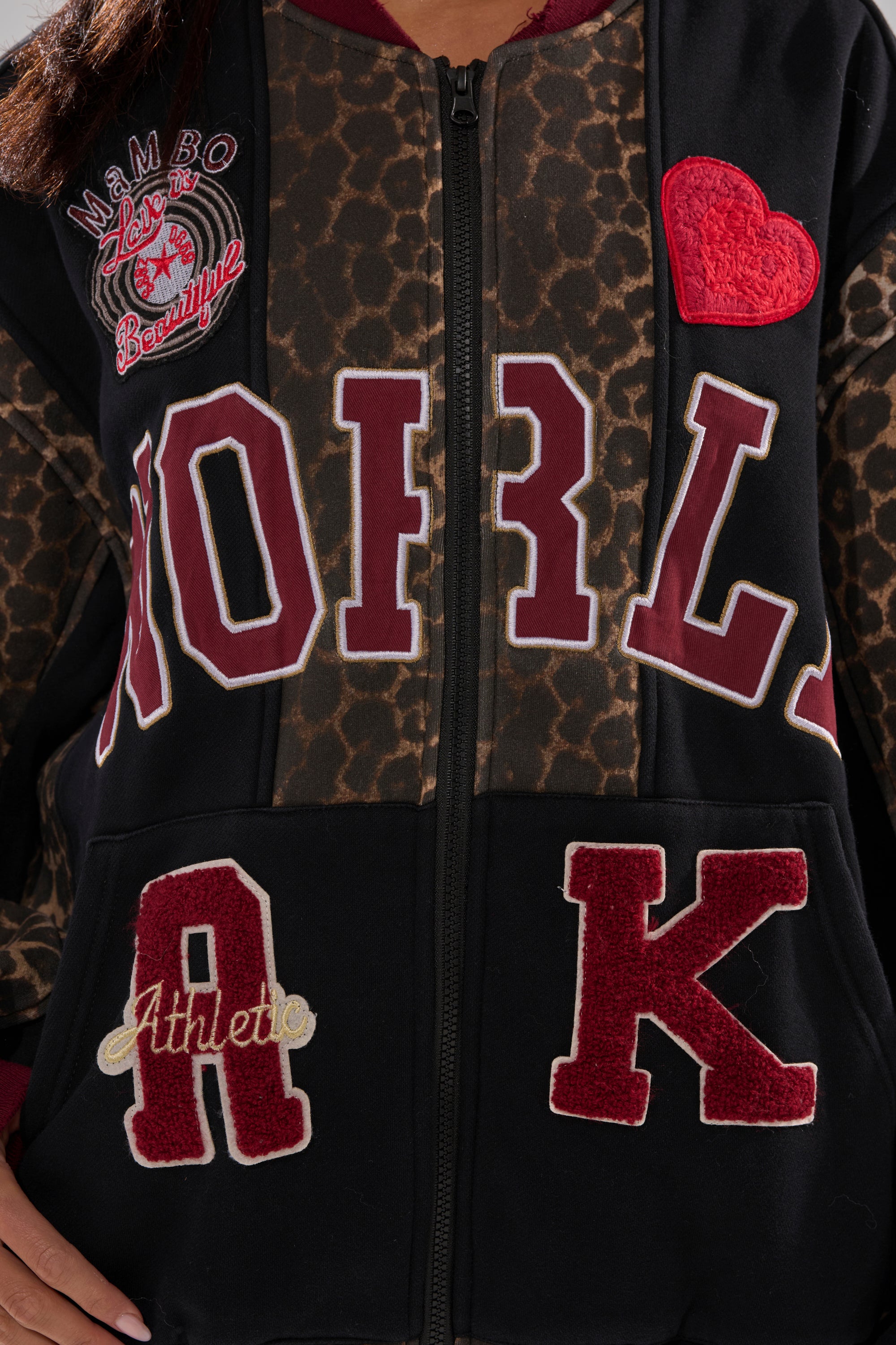 Close-up of the WORLD HOLD ON ZIP UP SWEATSHIRT, featuring varsity lettering, a red heart patch, "Athlete" text, leopard print accents, and "Mambo Beauty" embroidery on the left shoulder.