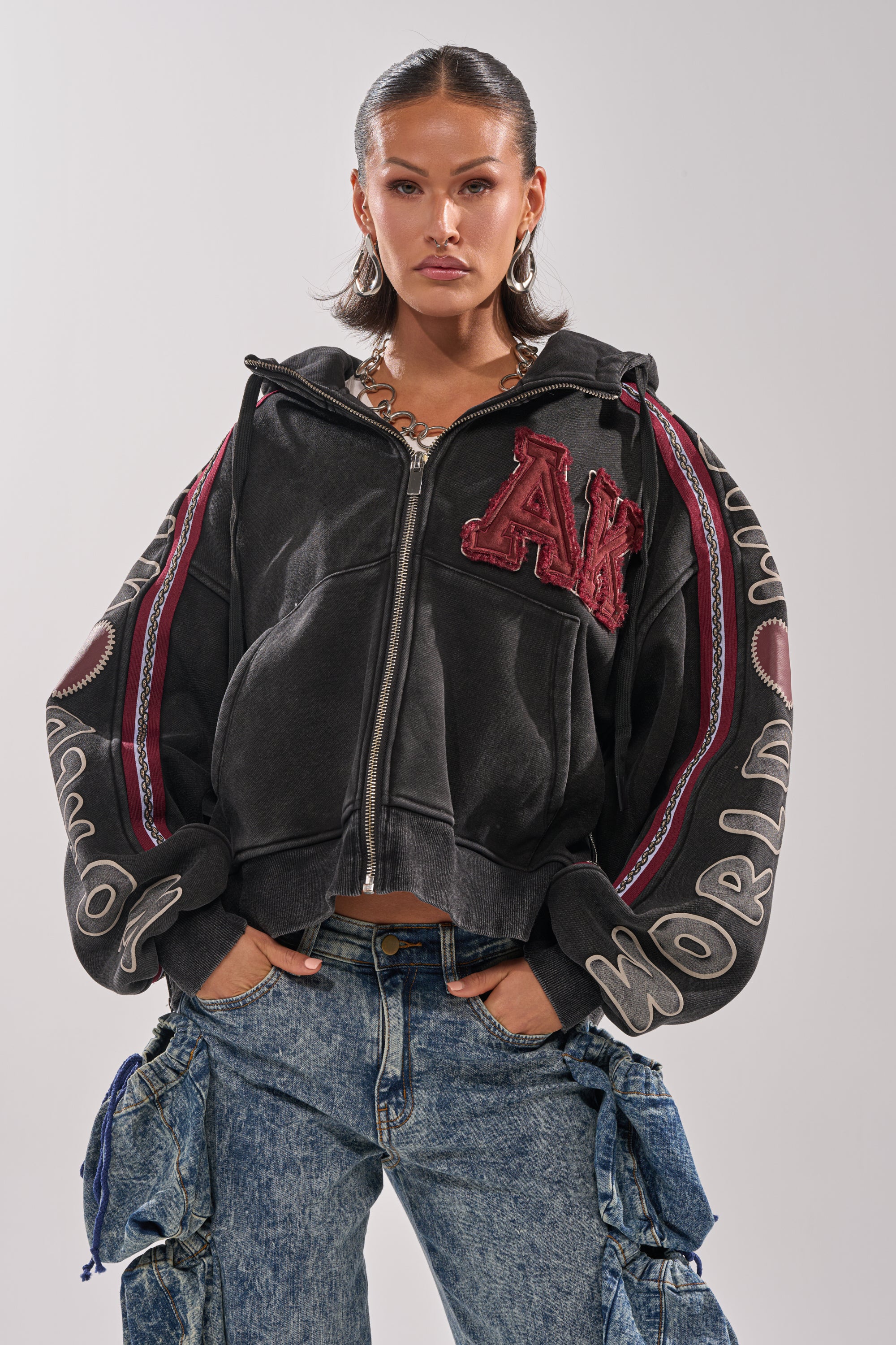 A woman exudes urban-chic style in the CITY LIVING GRAPHIC ZIP UP SWEATSHIRT, featuring bold patches and lettering, paired with loose blue cargo jeans. Her slicked-back hair and neutral expression complete the confident city look.