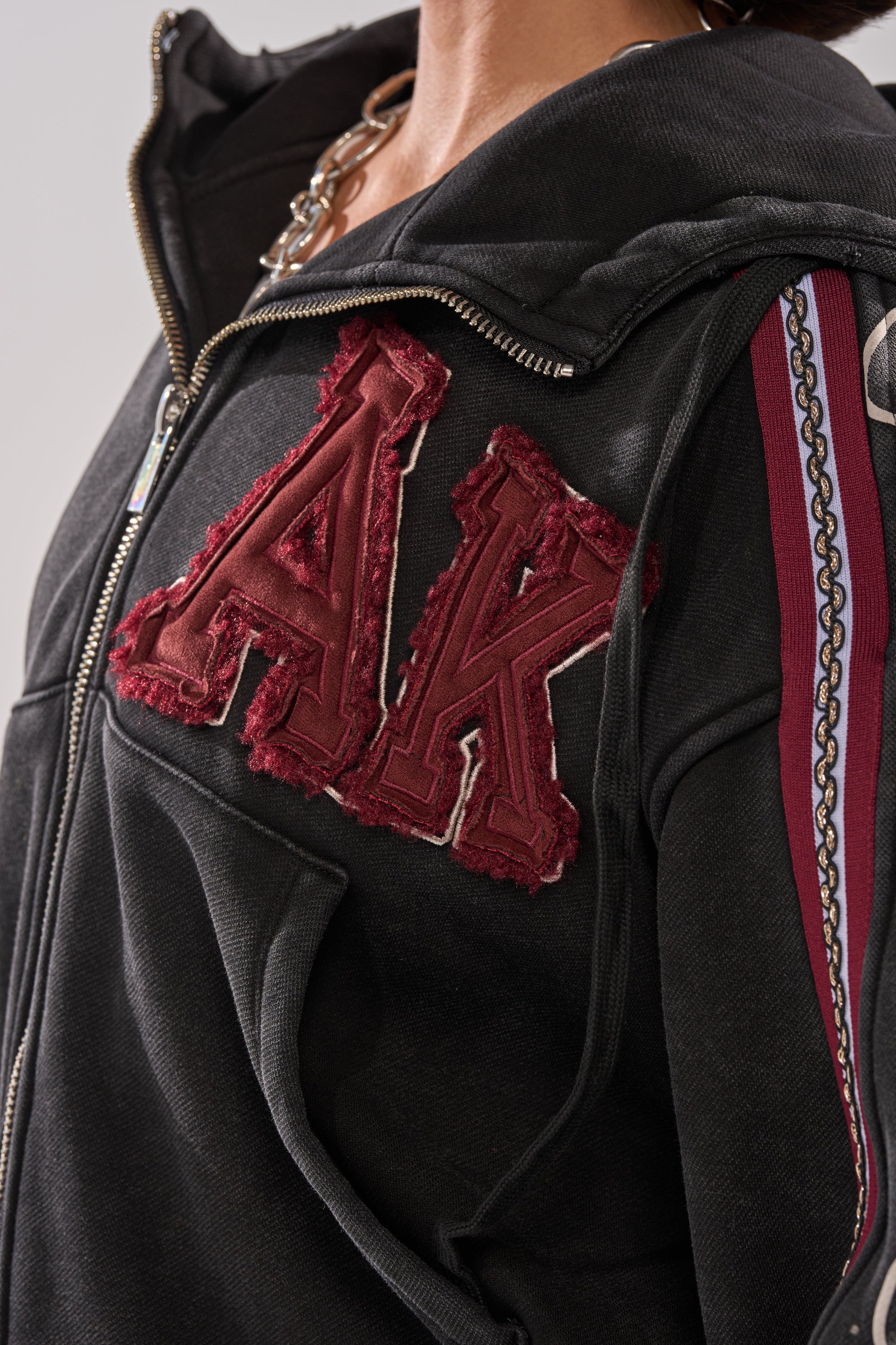 Close-up of a person wearing the CITY LIVING GRAPHIC ZIP UP SWEATSHIRT, an oversized hoodie with bold red textured "AK" letters on the chest and striped chain-patterned sleeves, ideal for an urban-chic look.