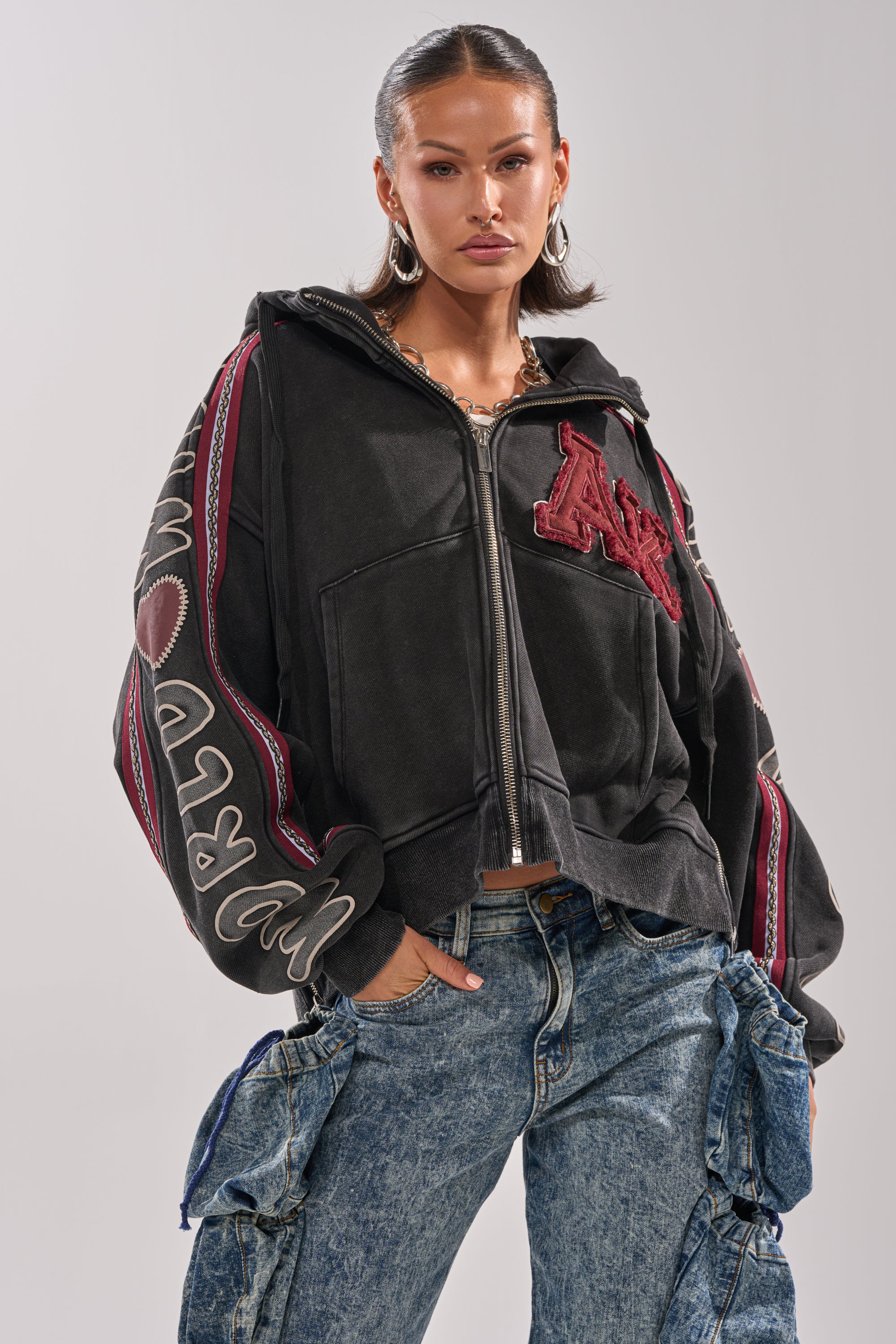 A woman poses confidently in the CITY LIVING GRAPHIC ZIP UP SWEATSHIRT, an urban-chic oversized hoodie with bold embroidered details, paired with baggy blue cargo jeans against a plain light background.