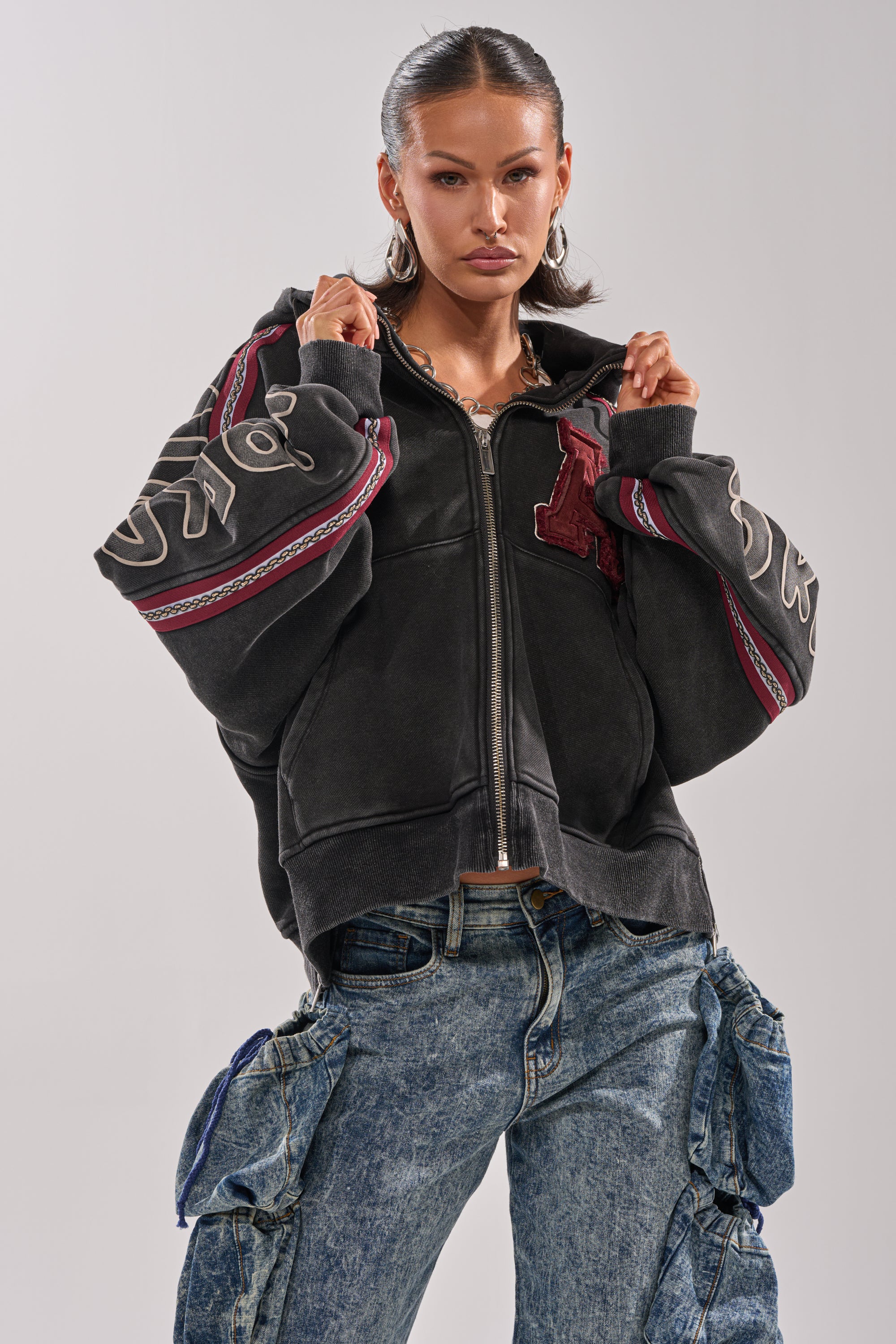 A woman stands confidently in a CITY LIVING GRAPHIC ZIP UP SWEATSHIRT with patterned sleeves and baggy blue cargo jeans, her hands on her oversized hoodie against a plain background.