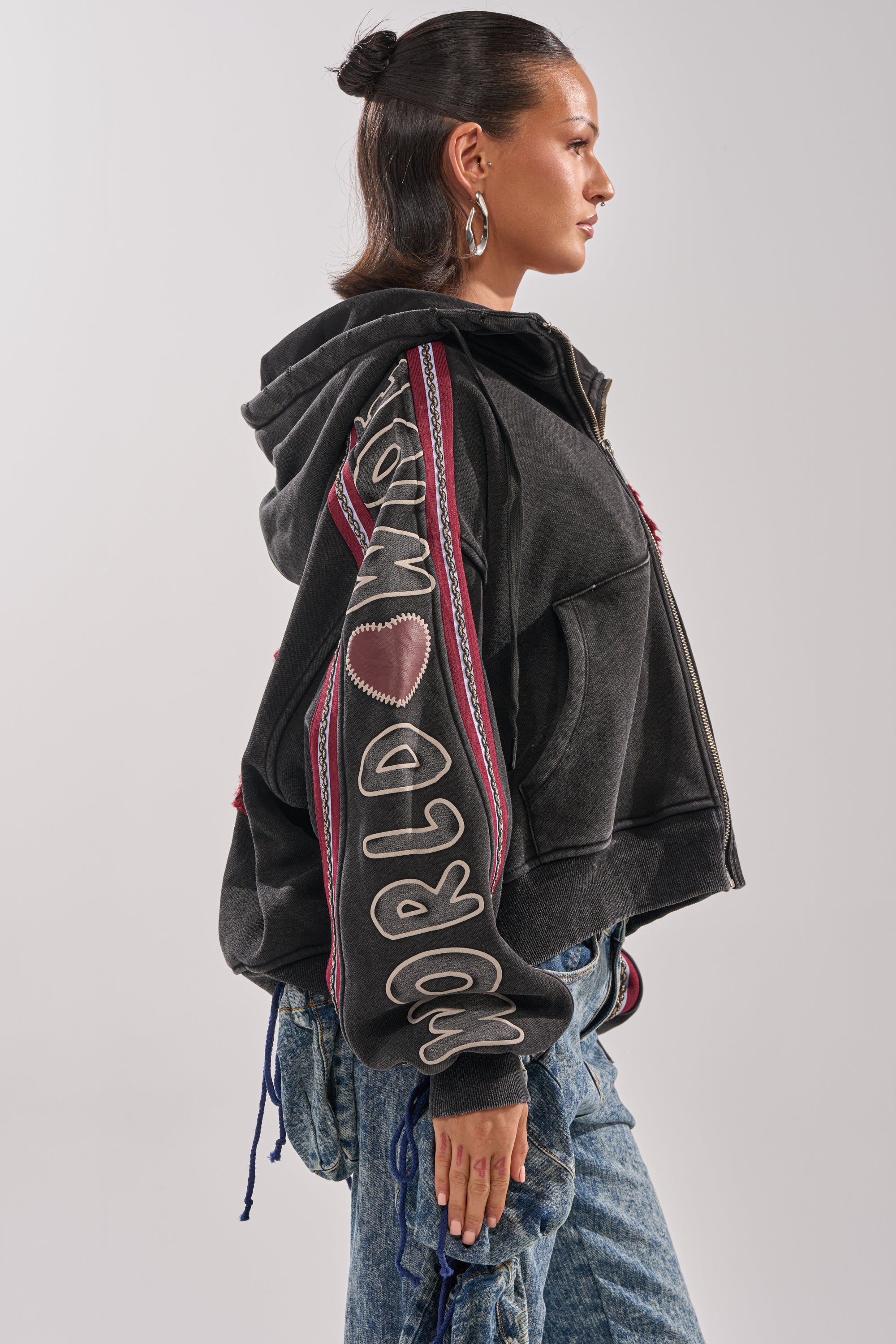 A woman in the CITY LIVING GRAPHIC ZIP UP SWEATSHIRT stands in profile, showing off a heart patch on her sleeve. She pairs it with blue jeans and large hoop earrings, her hair in a half-up bun for a relaxed urban look.