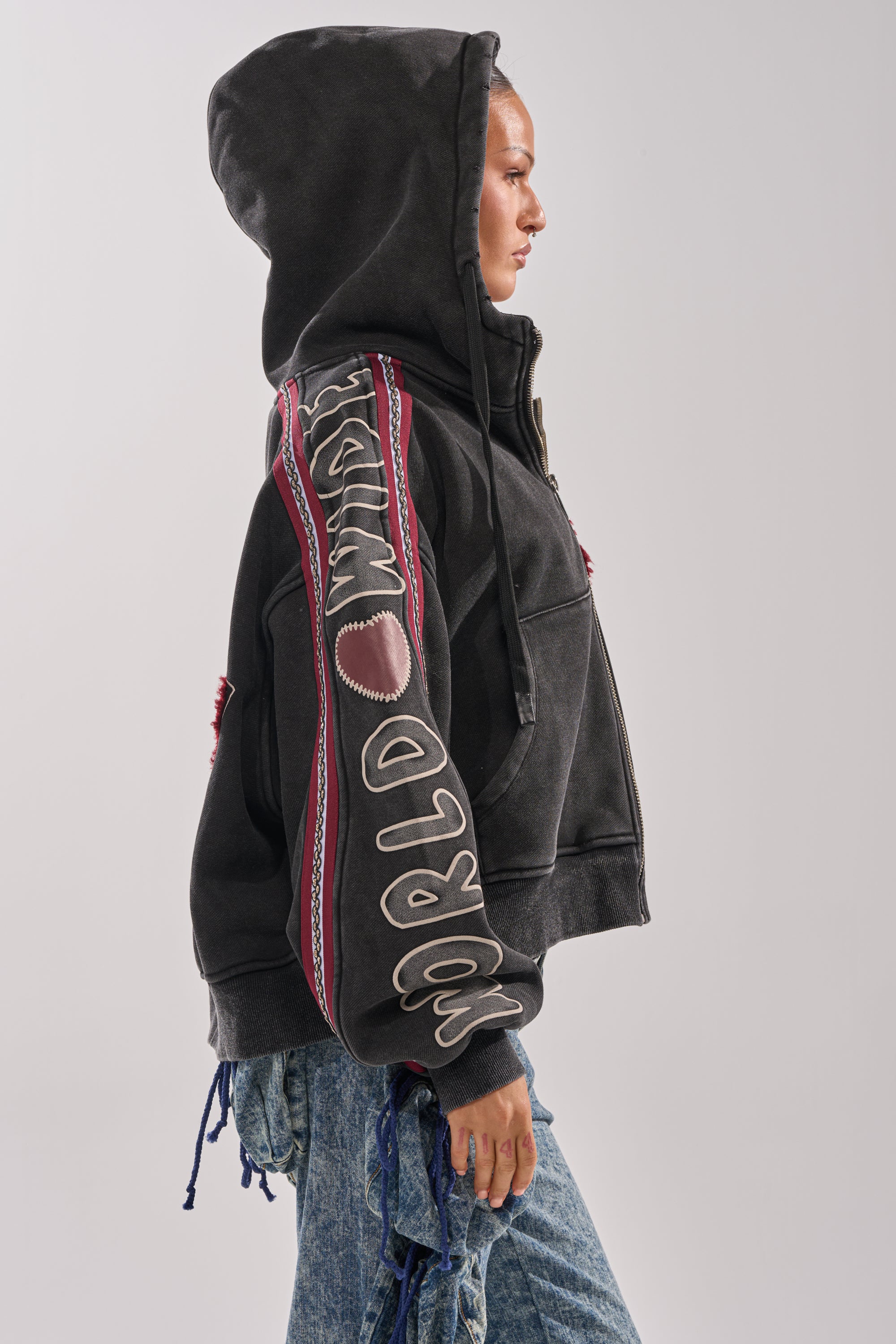Wearing the CITY LIVING GRAPHIC ZIP UP SWEATSHIRT with "WORLD WASTE" and sleeve stitching, paired with blue jeans, a person stands in profile against a plain light background.