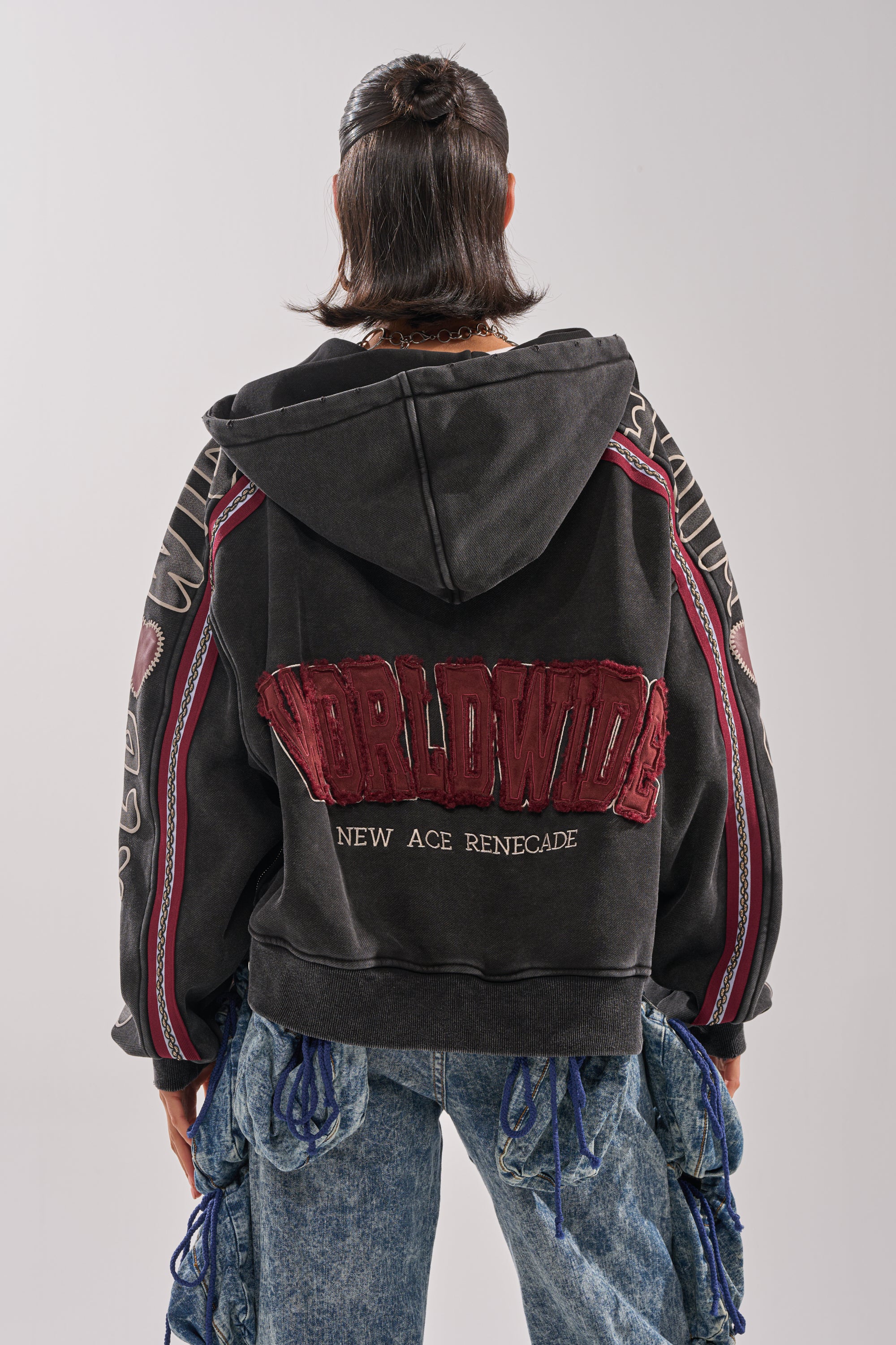 A person with dark, shoulder-length hair, seen from behind, wears the CITY LIVING GRAPHIC ZIP UP SWEATSHIRT—an oversized hoodie with "WORLDWIDE NEW AGE RENEGADE" in red on the back—and distressed blue jeans with blue fabric accents.
