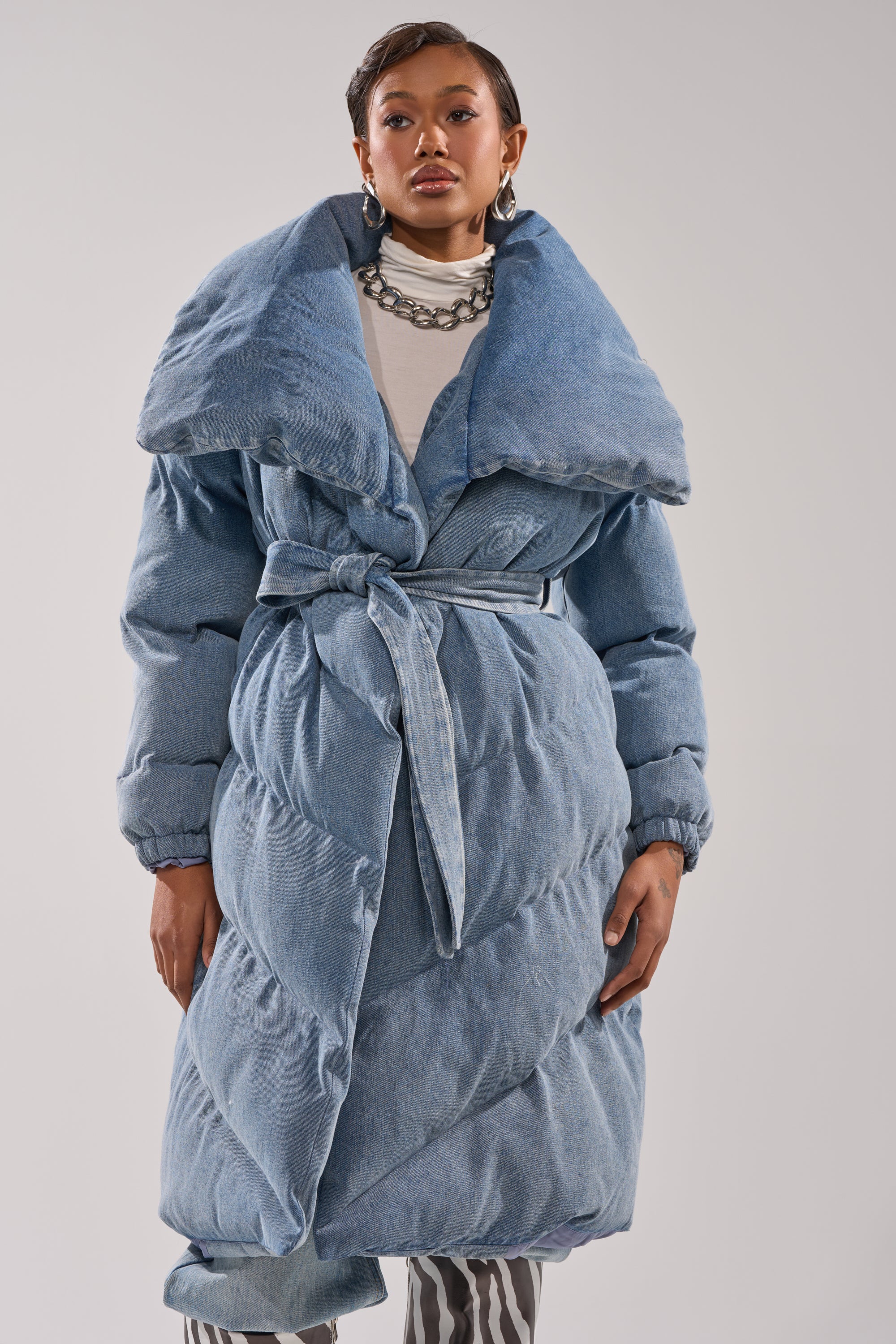 A woman models the I'M OUTSIDE DENIM PUFFER, a long light blue puffer coat with quilted padding and an oversized shawl collar, paired with a white turtleneck and chunky chain necklace against a plain background.