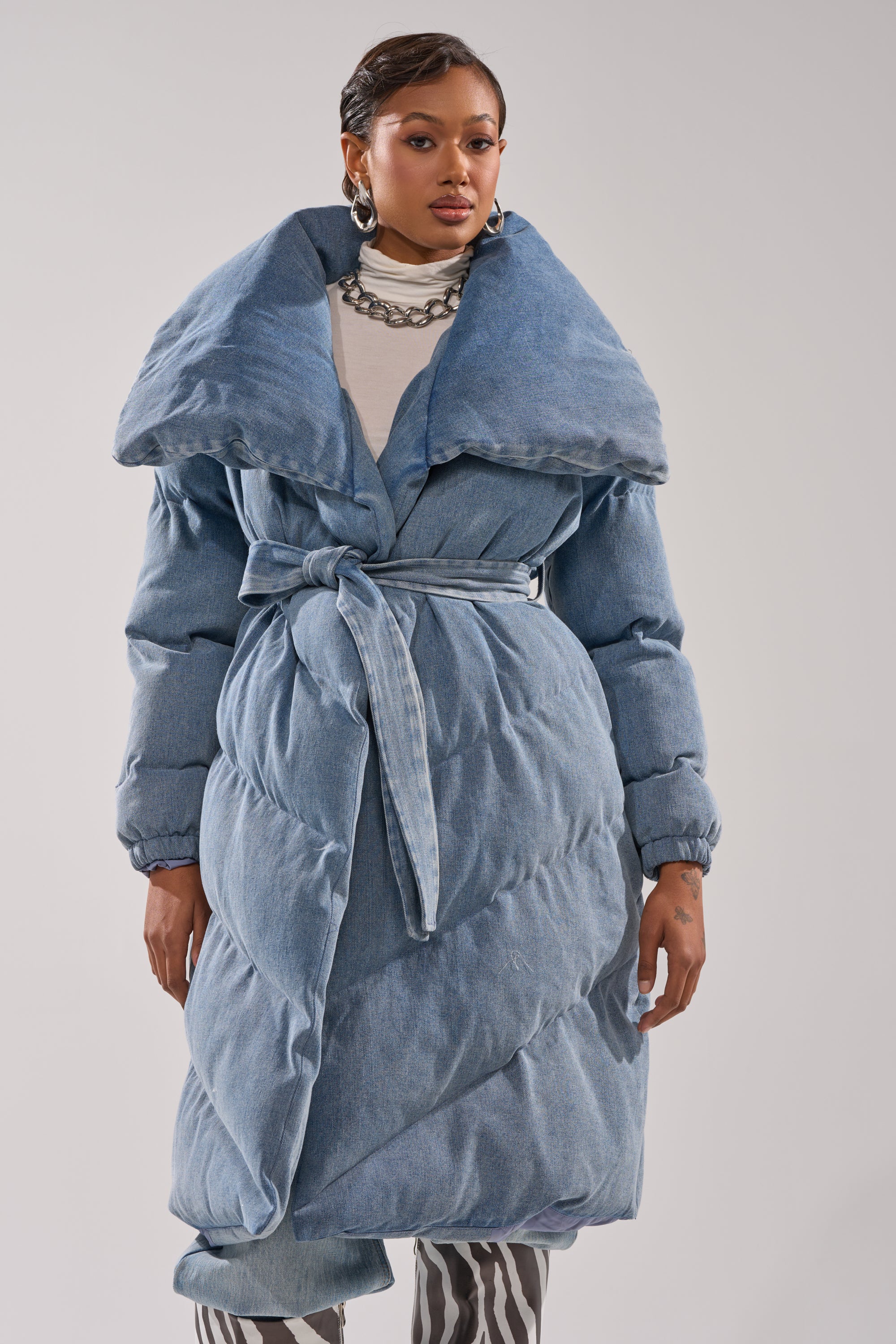 A woman wears the I'M OUTSIDE DENIM PUFFER, a light blue coat with an oversized shawl collar and belted waist, paired with a white shirt, chunky silver necklace, and patterned pants against a plain light background.