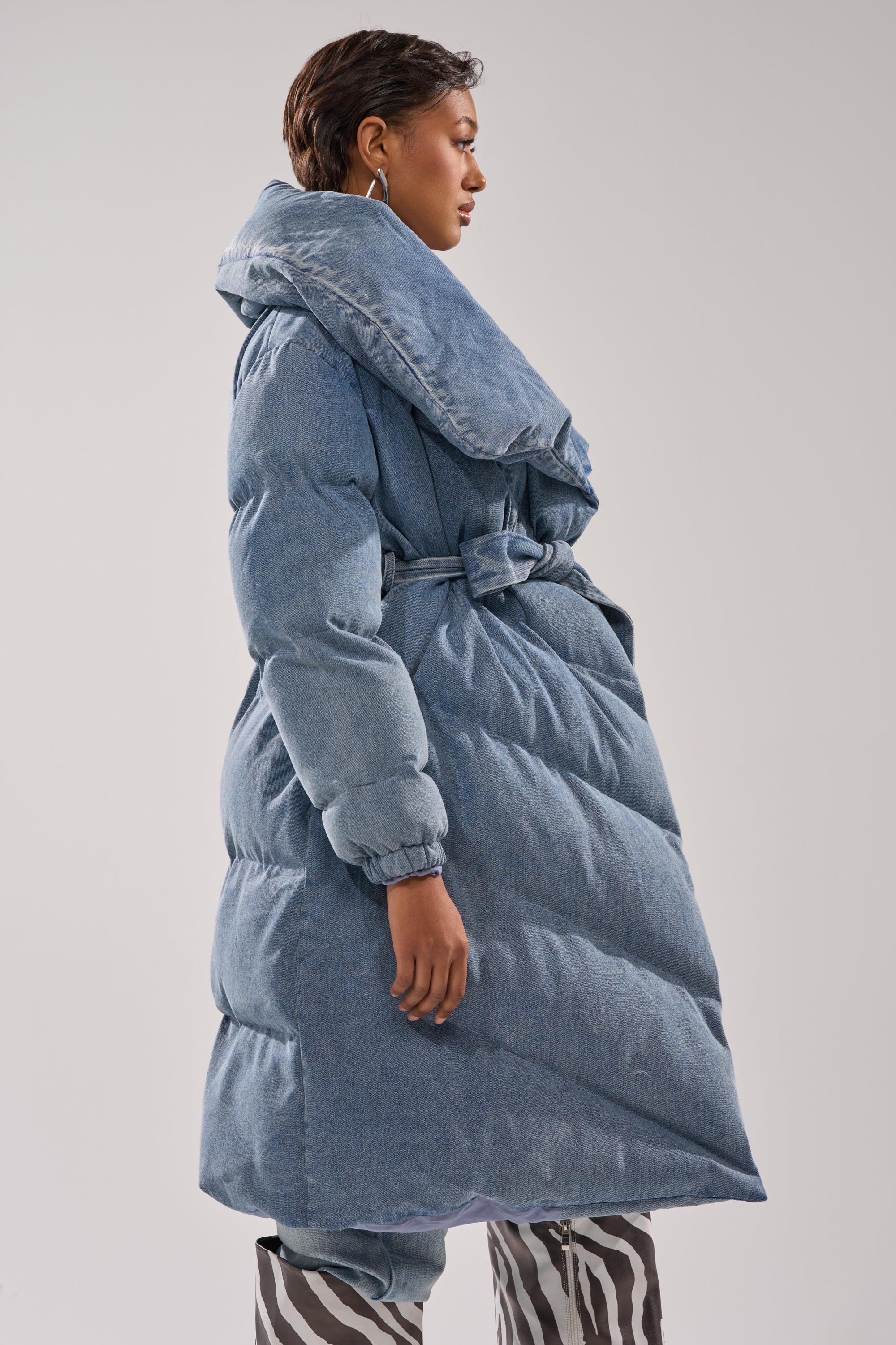 A woman in side profile poses in the I'M OUTSIDE DENIM PUFFER, a long blue quilted coat with an oversized shawl collar, styled with black-and-white striped pants and hoop earrings against a plain backdrop.
