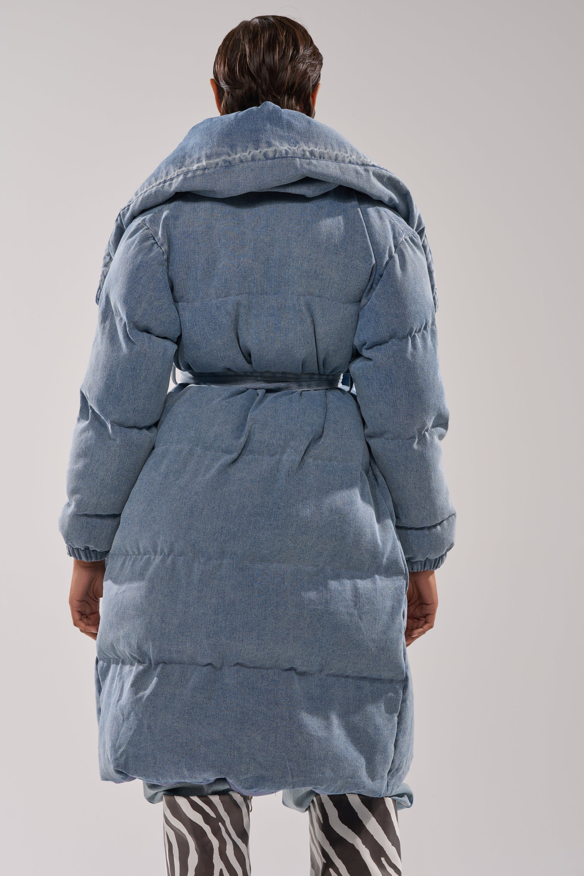 A person stands facing away, wearing the I'M OUTSIDE DENIM PUFFER—a light blue denim puffer coat with an oversized shawl collar and belted waist—paired with black and white zebra-print pants against a plain background.