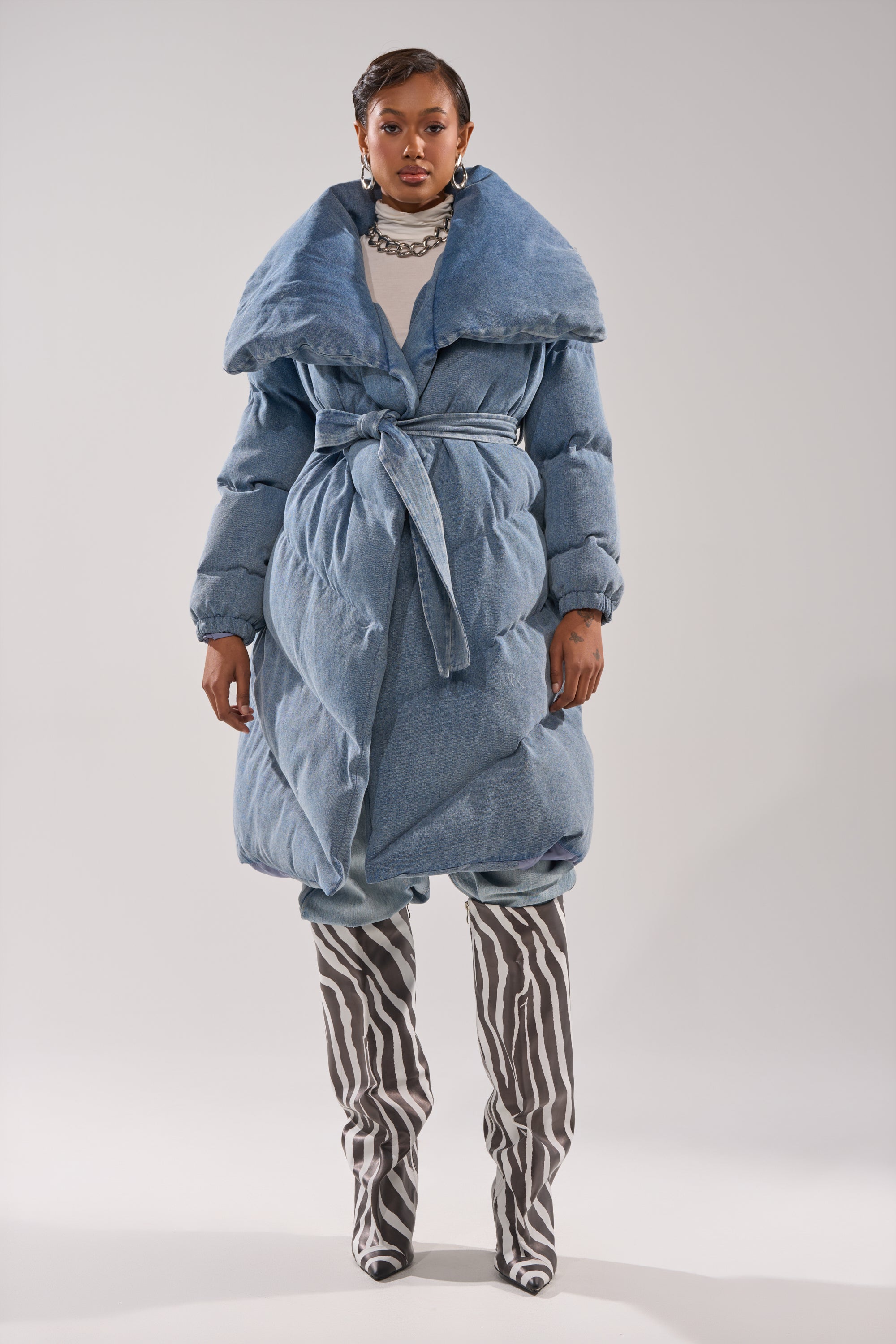 A woman faces forward in the I'M OUTSIDE DENIM PUFFER, a large light blue denim coat with an oversized shawl collar, layered over a white shirt and paired with knee-high black-and-white zebra-striped boots against a plain backdrop.