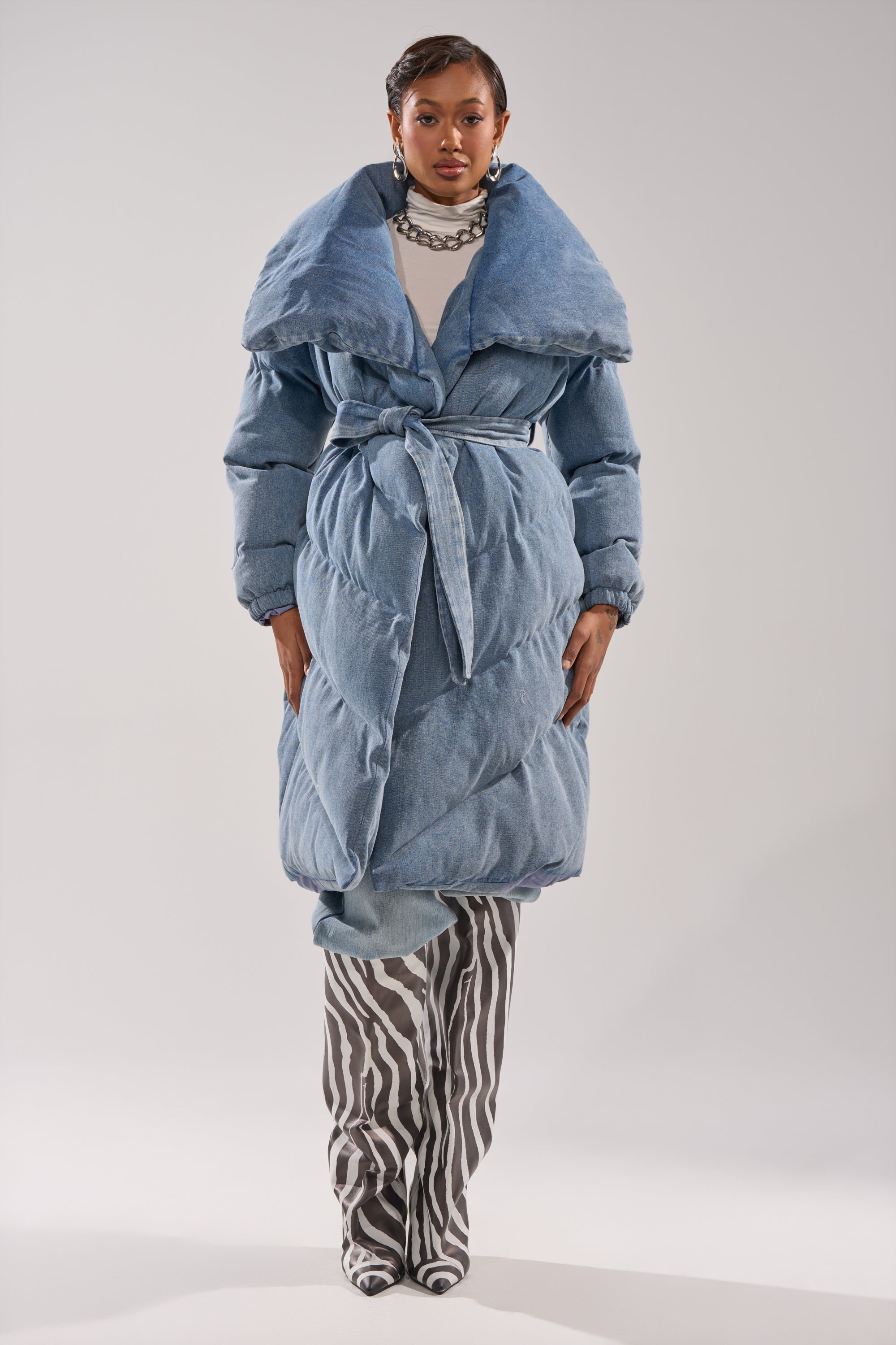 A person faces forward in the I'M OUTSIDE DENIM PUFFER, featuring an oversized shawl collar and a belted, wrap-style silhouette, paired with black-and-white zebra print pants against a plain, light background.