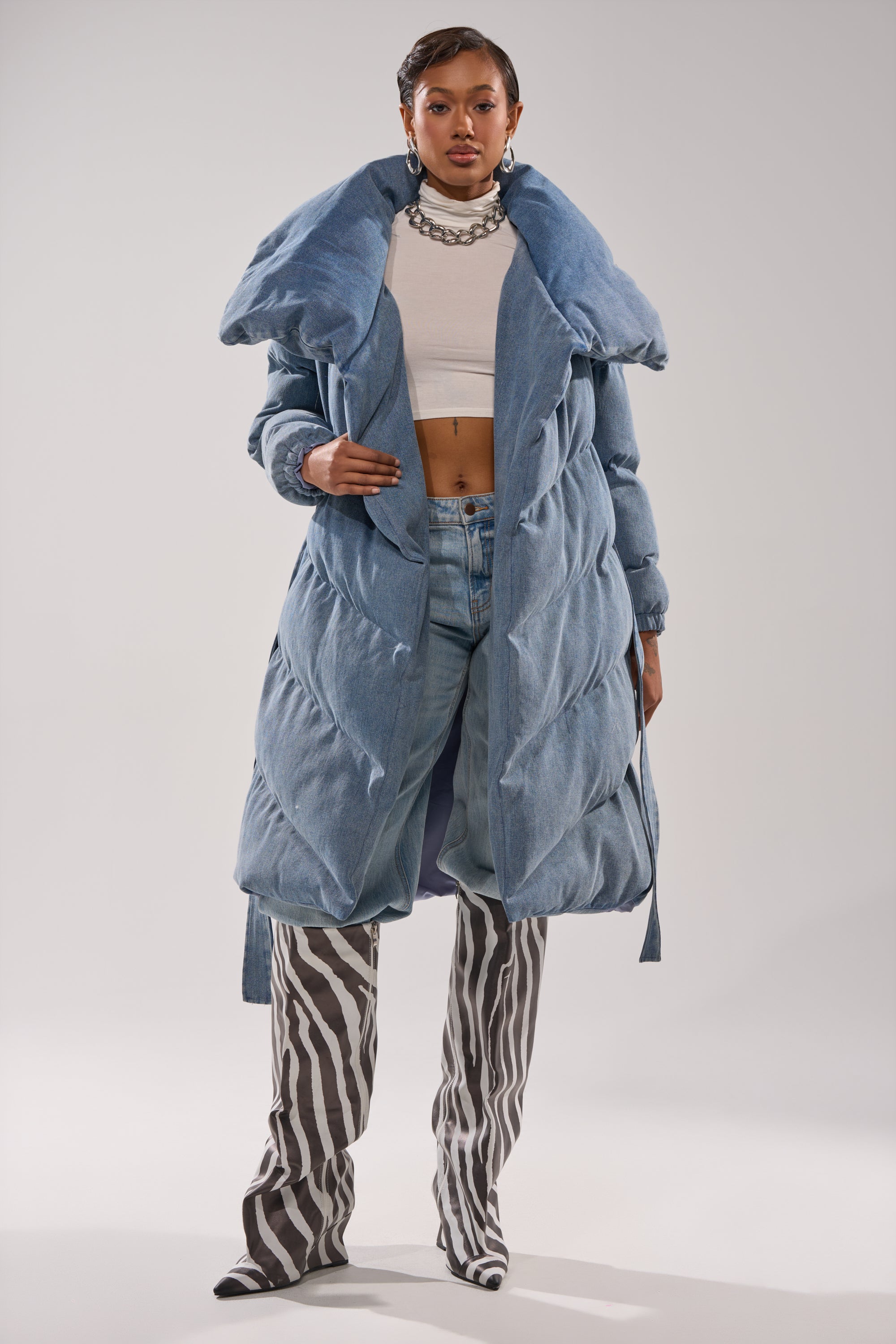 A woman poses confidently in the I'M OUTSIDE DENIM PUFFER, featuring a long blue design with an oversized shawl collar, styled with a white crop top, light blue jeans, chunky necklace, hoop earrings, and knee-high black-and-white zebra print boots.