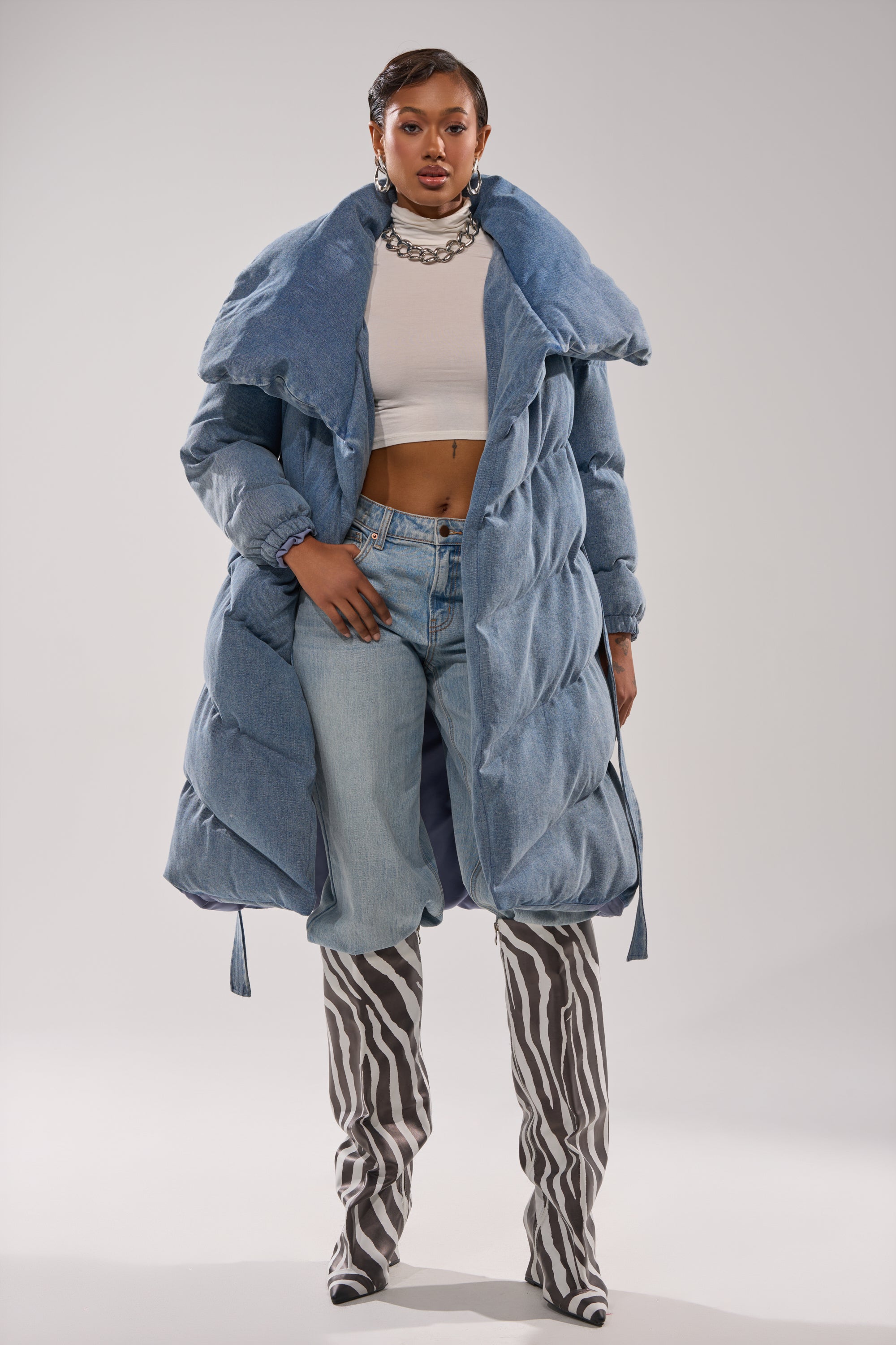 A woman stands confidently in the I'M OUTSIDE DENIM PUFFER with an oversized shawl collar, light blue jeans, a white crop top, chunky silver necklace, and zebra print knee-high boots against a plain light background.