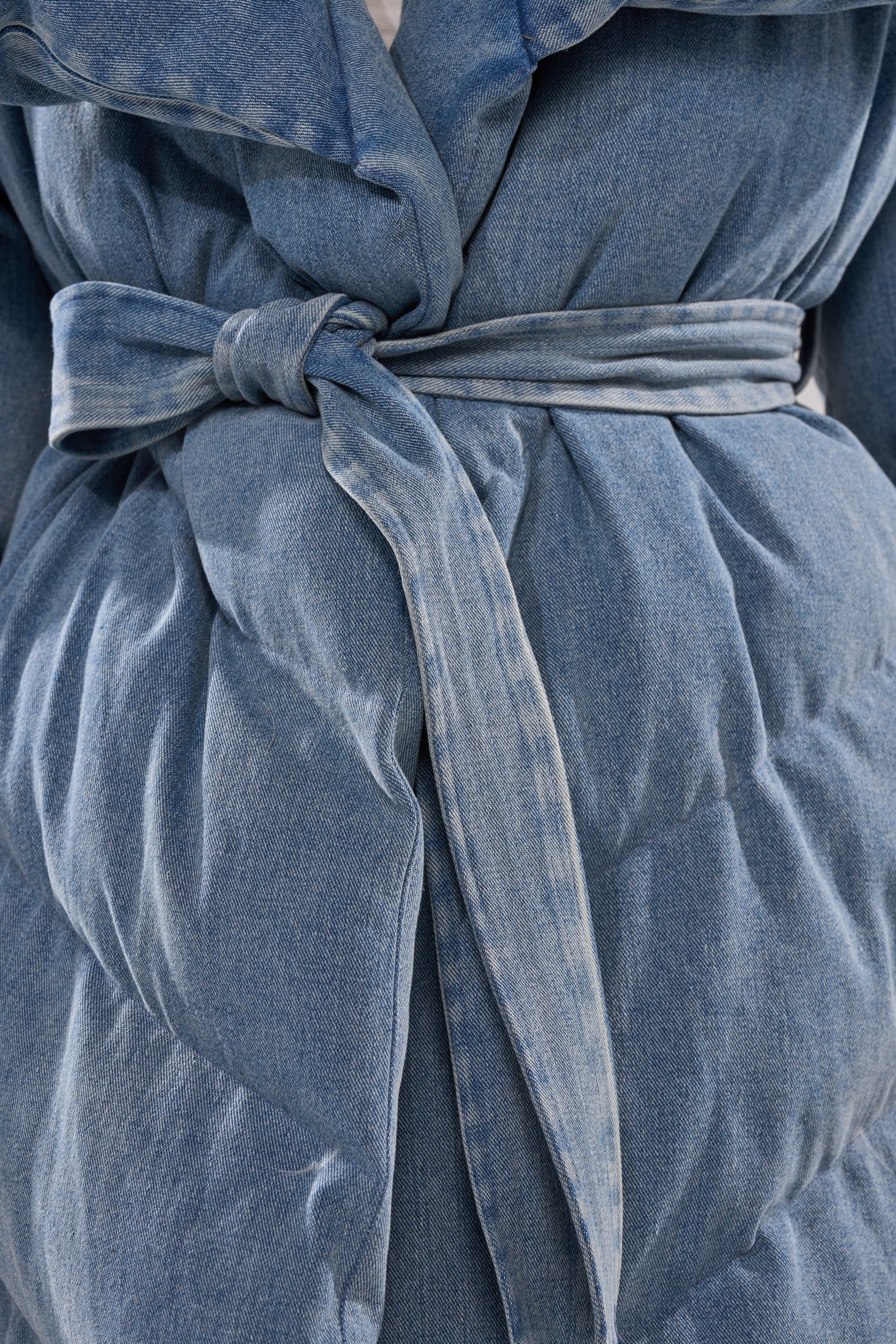 Close-up of someone wearing the I'M OUTSIDE DENIM PUFFER, featuring an oversized shawl collar, tied waist belt, and textured quilted blue denim fabric.