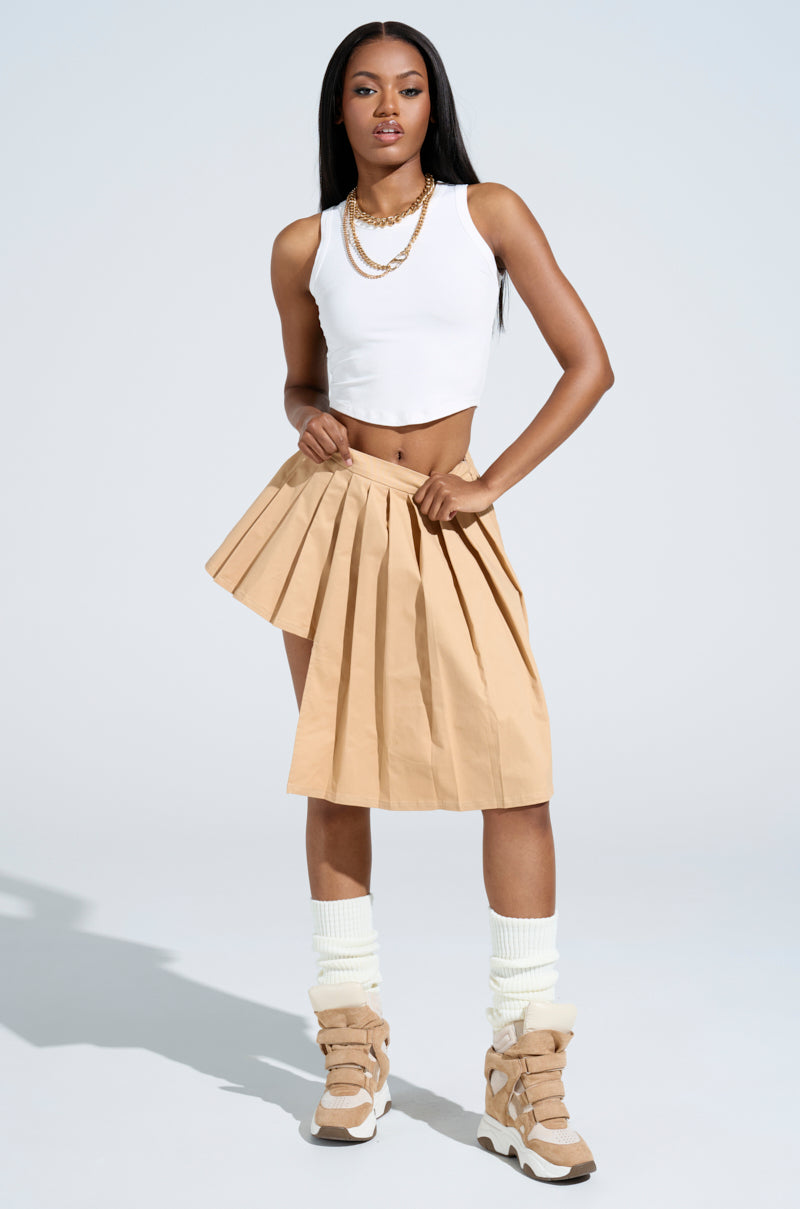 A woman exudes confidence in a white crop top and the SHARI BABY PLEATED MINI SKIRT, featuring an asymmetrical hem. She pairs her outfit with layered necklaces, beige boots, and white leg warmers against a simple light gray background that accentuates her chic look.