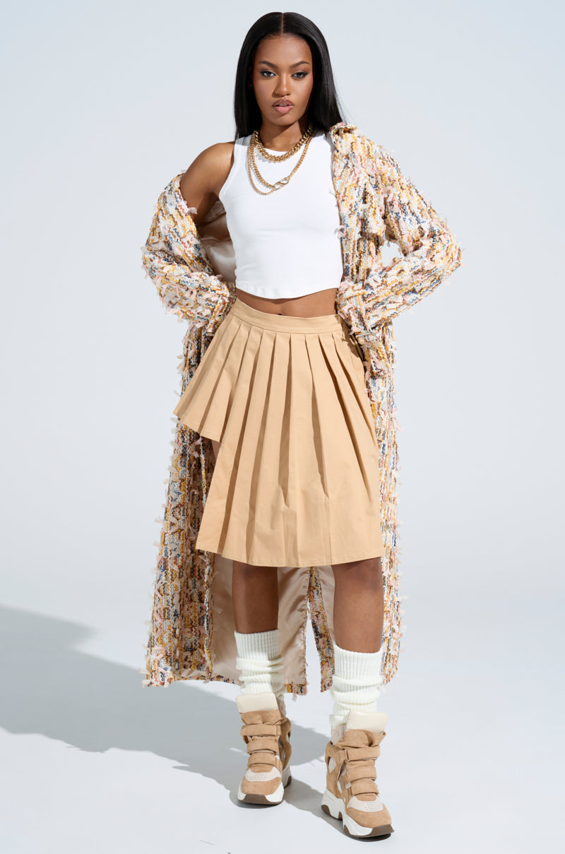 A woman confidently poses in a white crop top, layered gold necklaces, a long textured coat, and the SHARI BABY PLEATED MINI SKIRT in beige, complementing her beige boots with white socks. The plain background highlights her modern ensemble's asymmetrical hem and chic style.