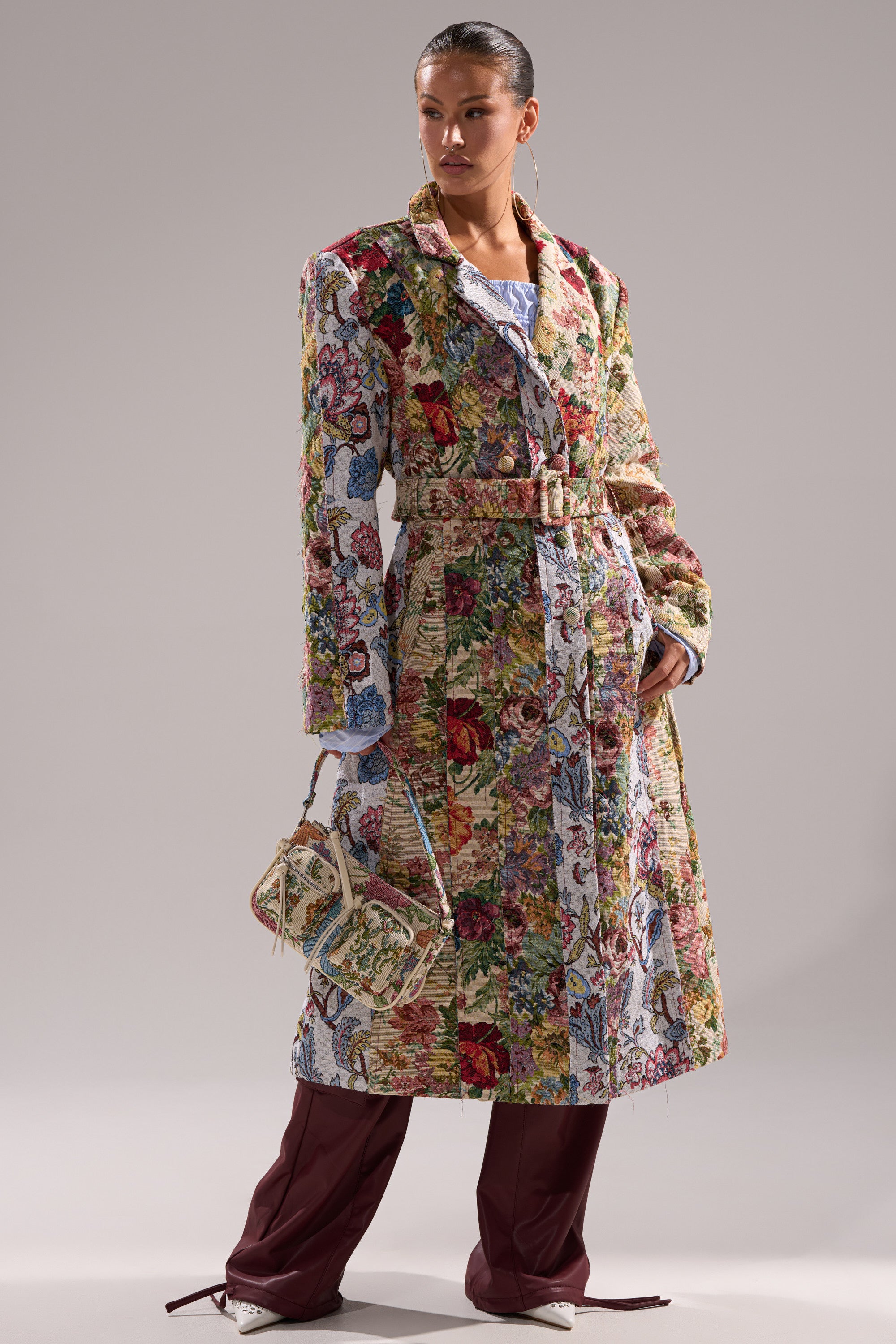 A person stands confidently in the SUNDAY PATCHWORK TRENCH, a colorful floral jacquard coat, paired with a matching handbag, wide-legged maroon pants, and white shoes against a plain background. 0