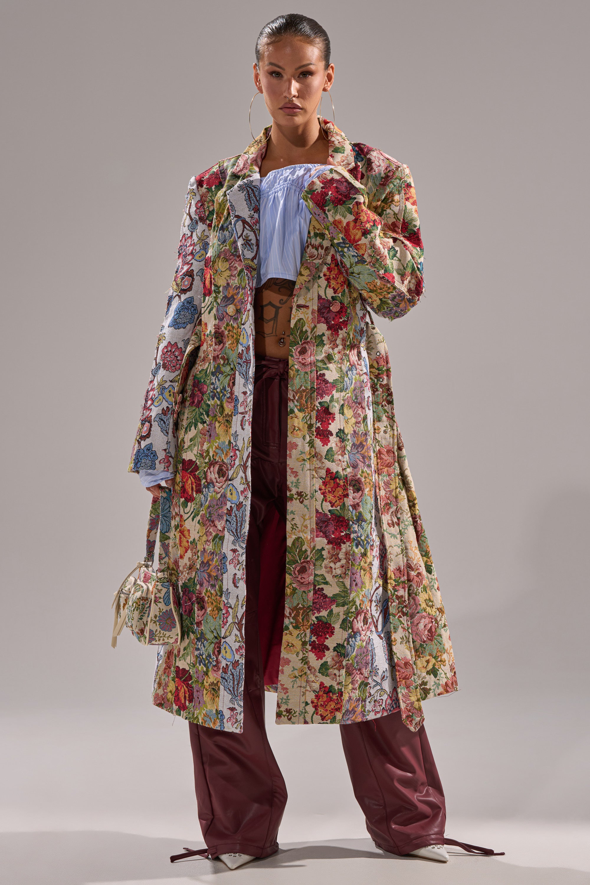 A person models the SUNDAY PATCHWORK TRENCH over a blue top and maroon wide-leg pants, paired with light shoes and a small patterned bag, standing against a plain gray background. 1