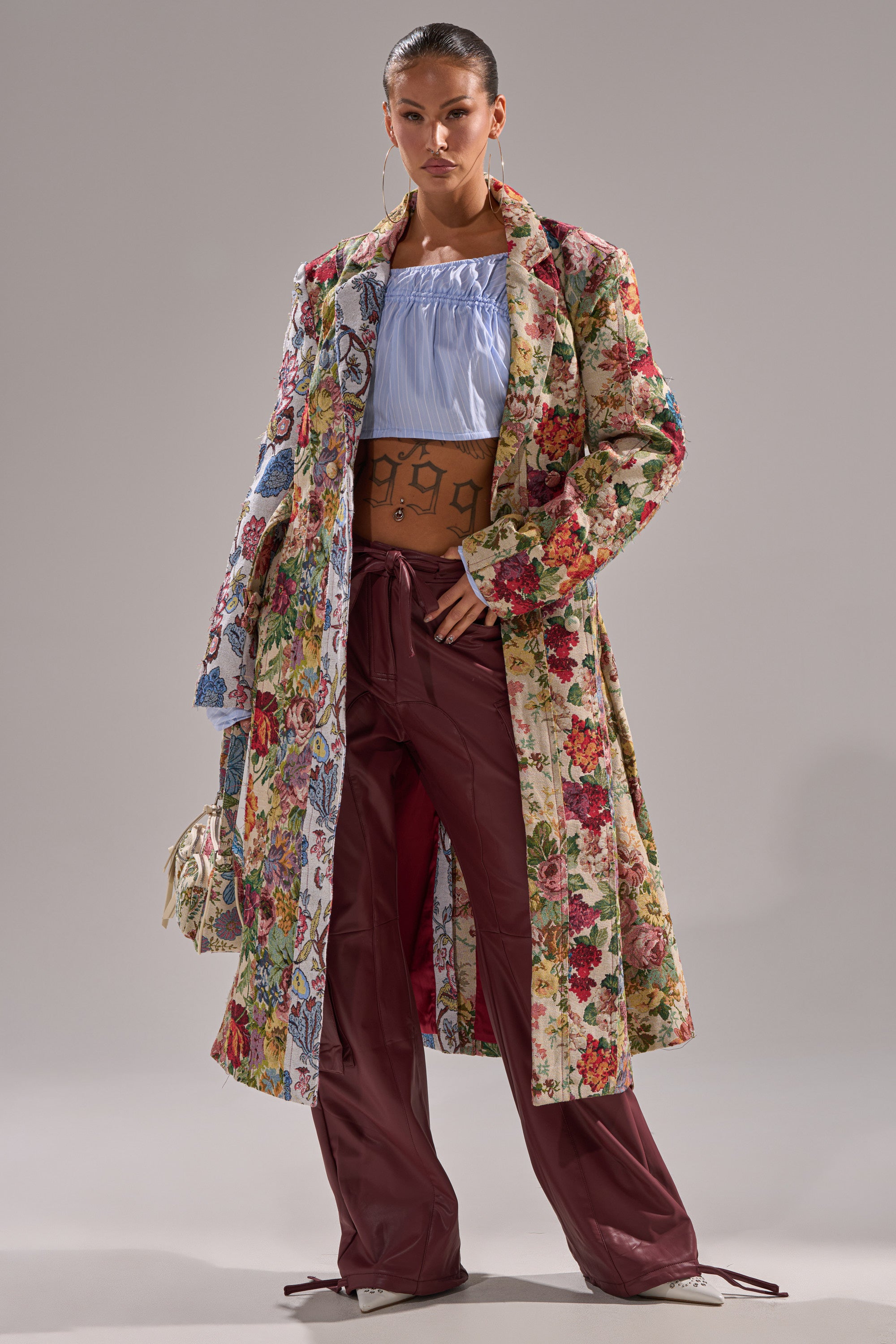 Wearing the SUNDAY PATCHWORK TRENCH over a light blue off-shoulder crop top and loose maroon pants, a confident woman holds a small handbag while posing against a plain background. 3