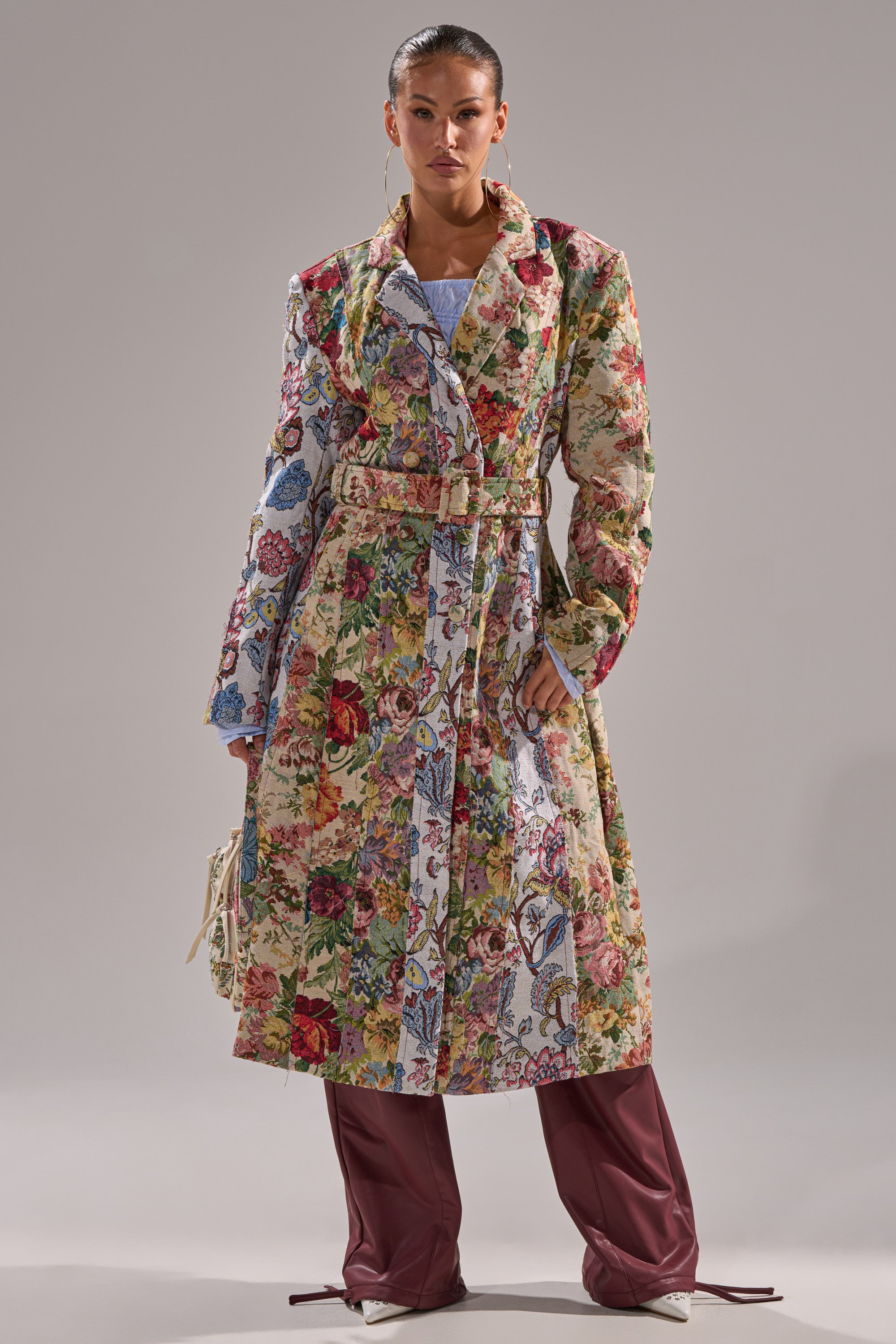 A woman faces forward in the SUNDAY PATCHWORK TRENCH, featuring a colorful floral design, paired with maroon wide-leg pants. She holds a small bag, with slicked-back hair, posing confidently against a plain background. 4