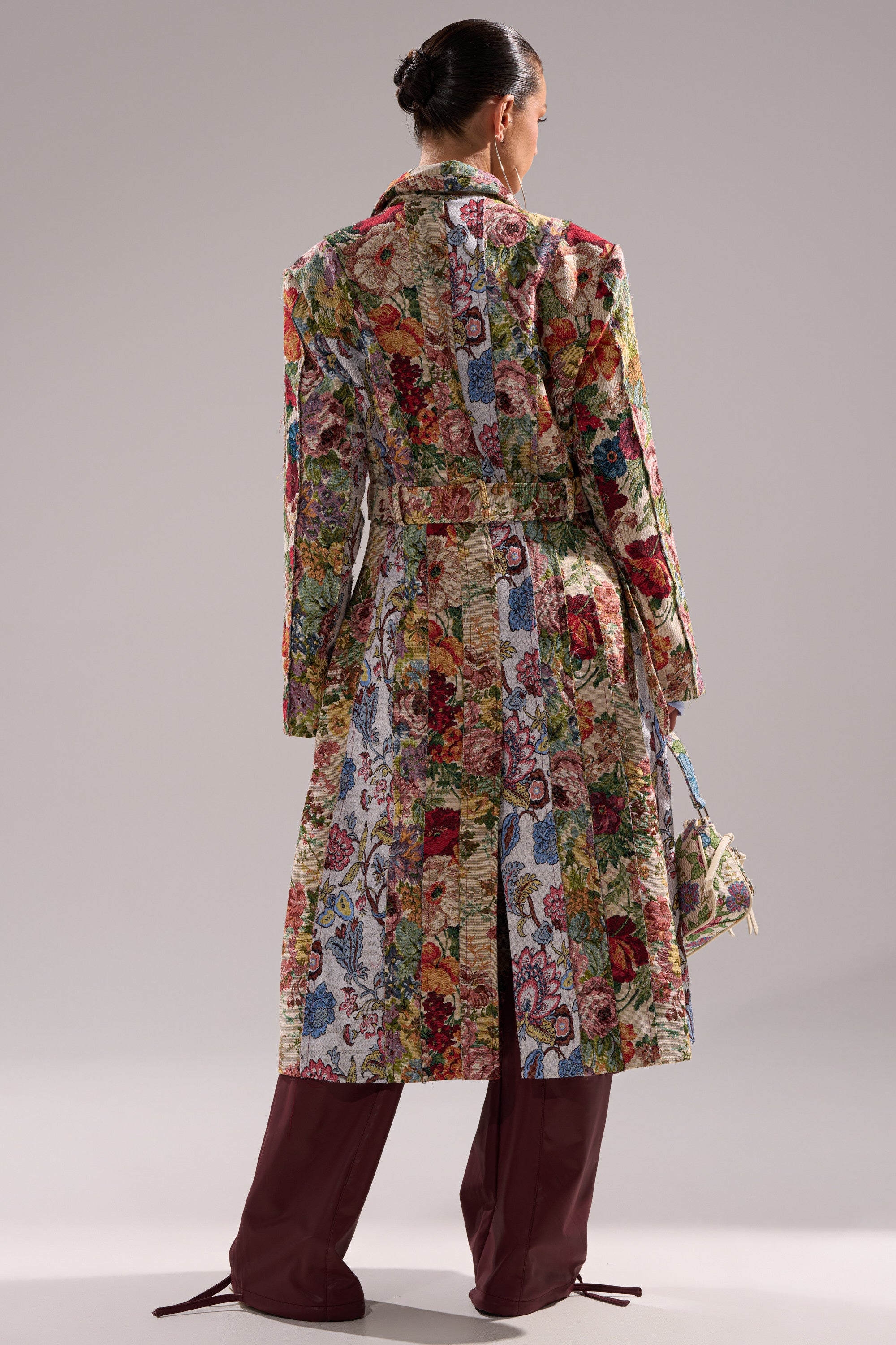 A person stands facing away, wearing the SUNDAY PATCHWORK TRENCH and burgundy knee-high boots, holding a small bag. Their hair is styled in a neat bun, set against a neutral background. 6