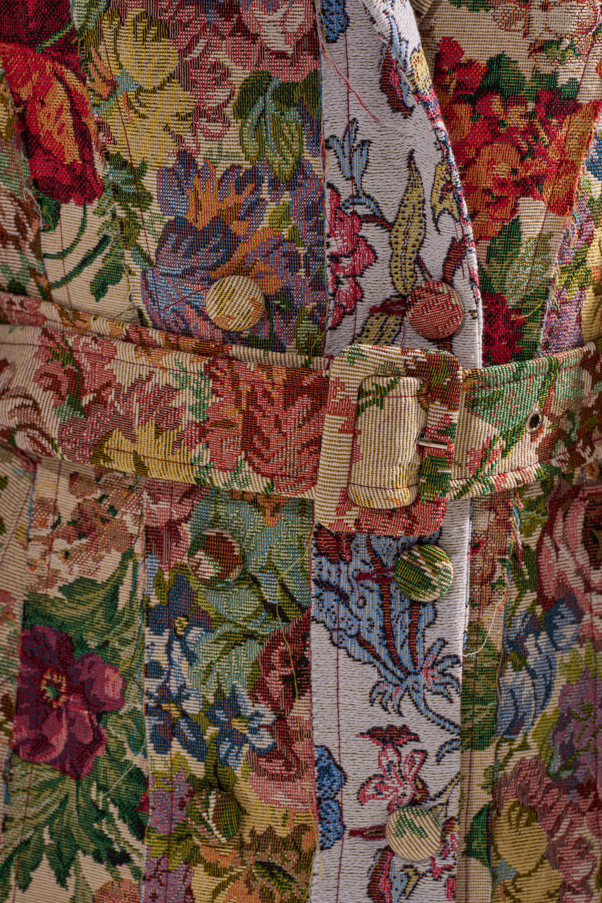 Close-up of the SUNDAY PATCHWORK TRENCH shows its vibrant floral jacquard fabric, detailed woven flower patterns, and a matching fabric belt with buckle, highlighting the rich texture and intricate design. 7