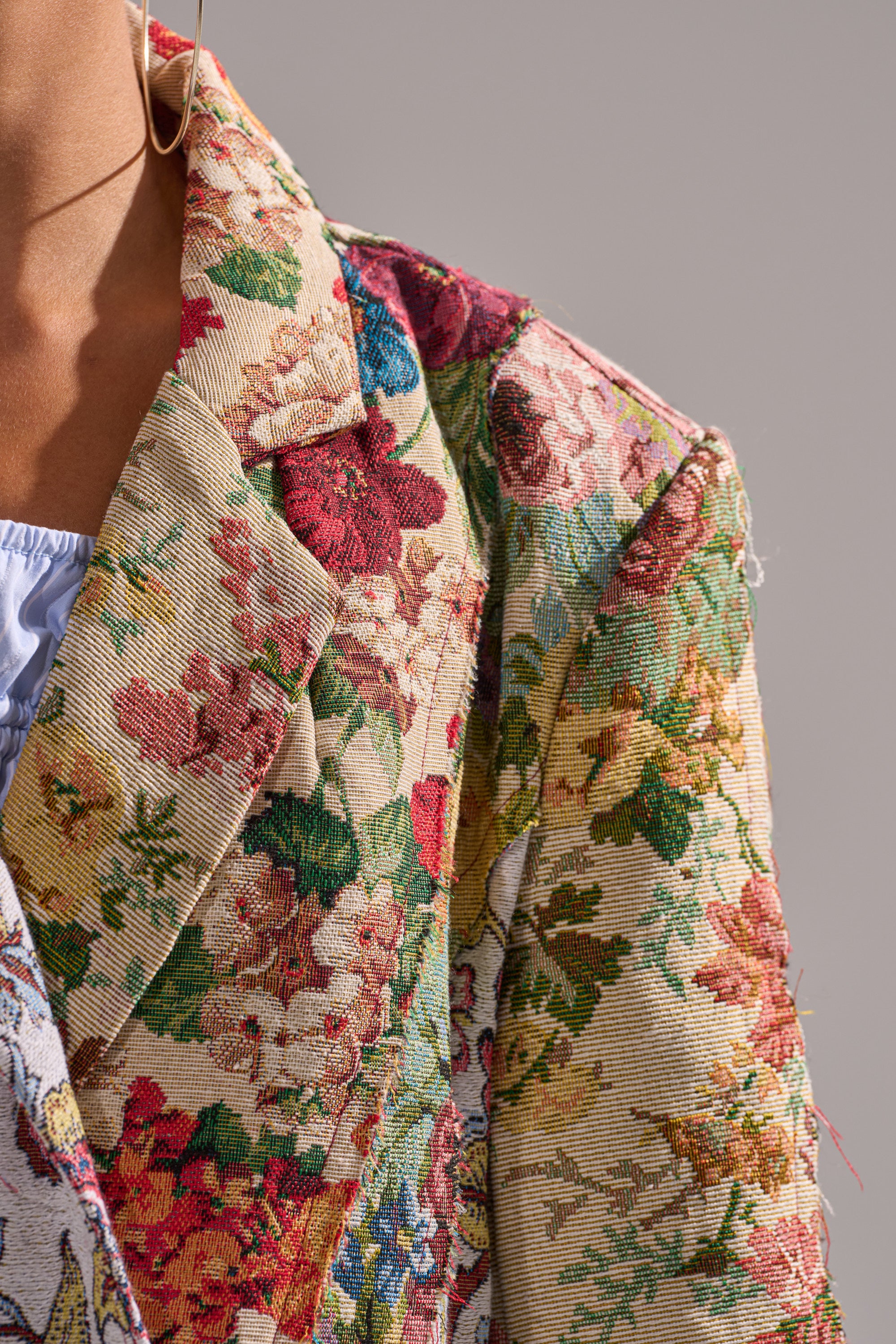 Close-up of a person's shoulder wearing the SUNDAY PATCHWORK TRENCH, a colorful floral jacquard jacket with textured fabric. Part of a blue shirt and an earring are also visible against a neutral background. 8