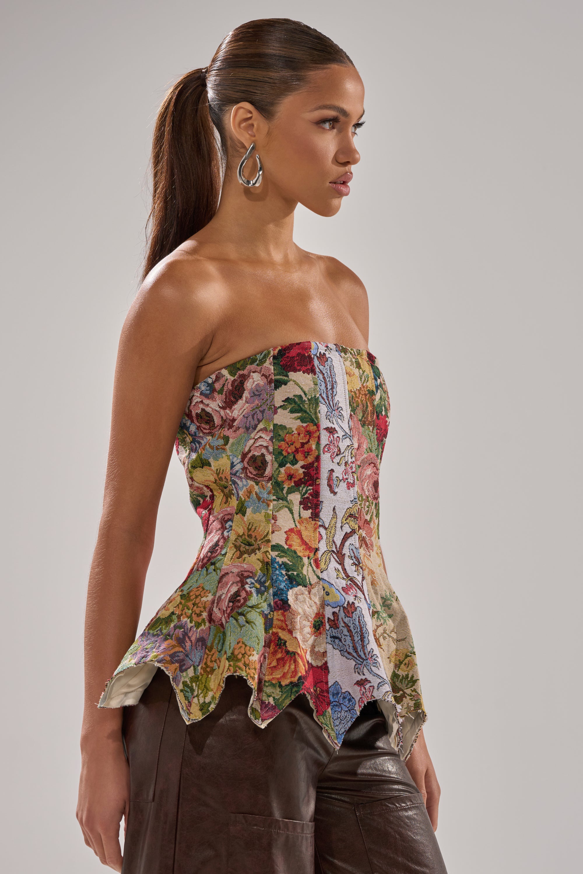 A woman with long brown hair in a ponytail wears the SUNDAY PATCHWORK CORSET, a strapless tapestry-style top with peplum detail, paired with dark pants. She stands sideways against a plain gray background.