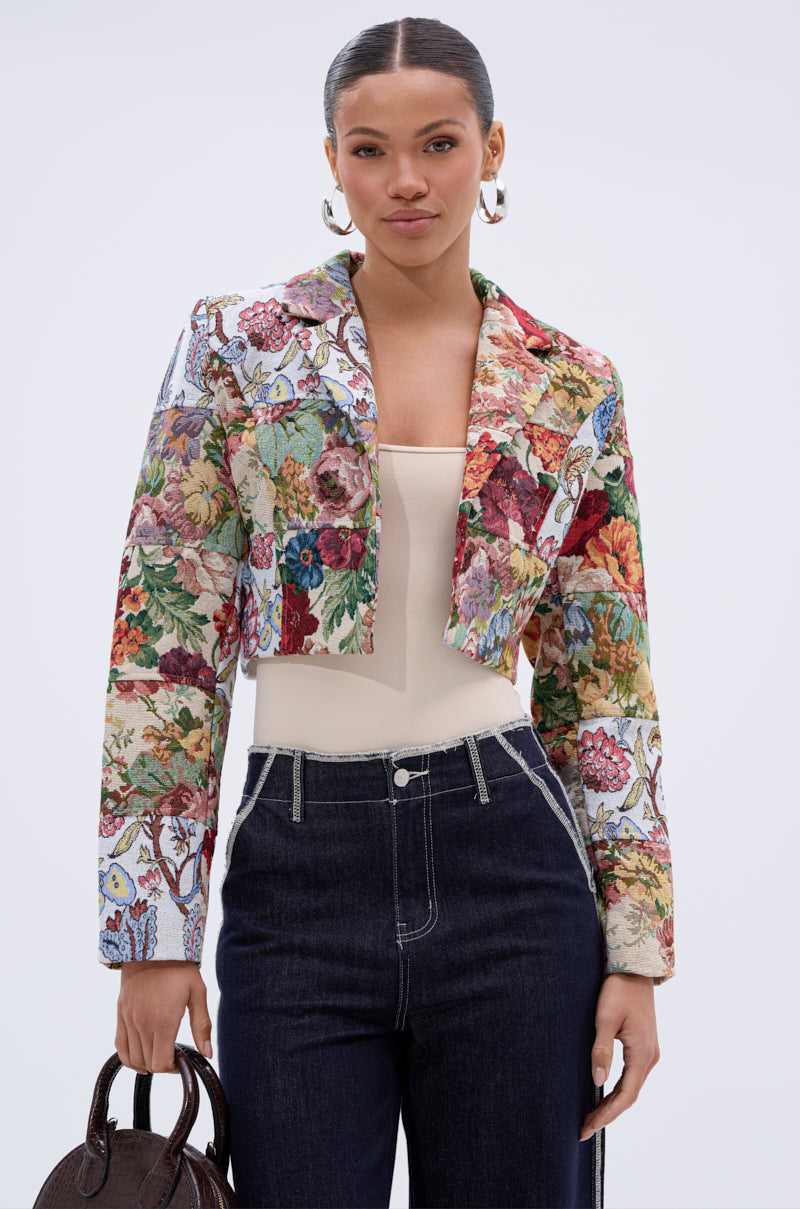 A woman exudes confidence in the TAKE A HINT PATCHWORK BLAZER, styled over a beige top and dark high-waisted jeans. She carries a brown handbag, wears large hoop earrings, and stands against a plain light background.