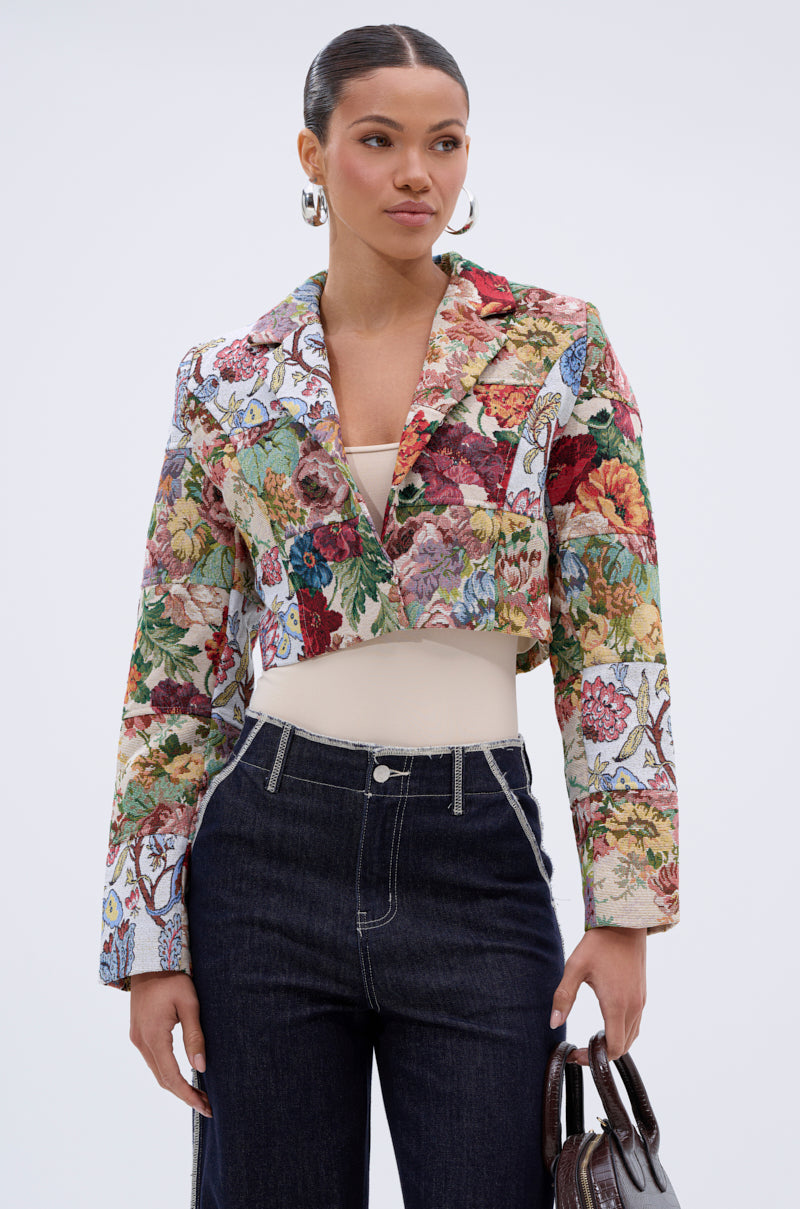 A woman wears the TAKE A HINT PATCHWORK BLAZER—a colorful, floral-patterned vintage jacquard piece—over a beige top and dark high-waisted jeans, holding a brown handbag against a plain light background.