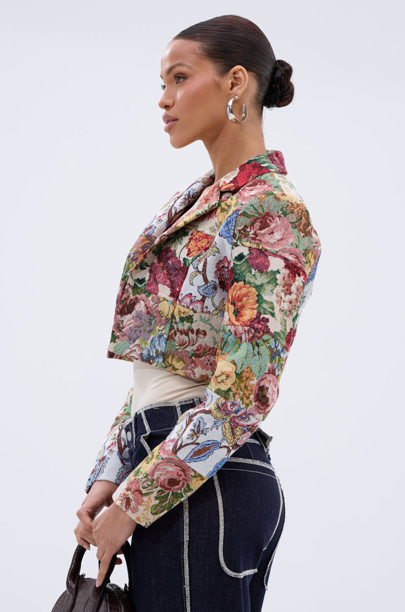 A woman in profile wears the TAKE A HINT PATCHWORK BLAZER with bold florals, paired with large hoop earrings, dark jeans featuring white stitching, and carries a small brown handbag against a plain white background.
