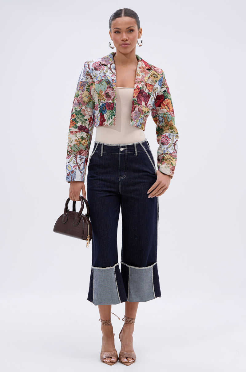 A woman faces forward in the TAKE A HINT PATCHWORK BLAZER, a beige top, dark cropped wide-leg jeans with exposed seams, strappy heels, hoop earrings, and a small brown handbag.