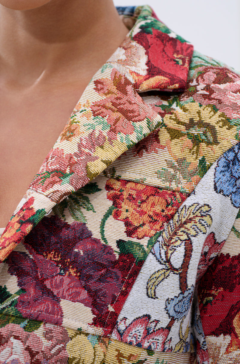 A close-up of a person wearing the TAKE A HINT PATCHWORK BLAZER, showcasing its woven texture and colorful floral pattern with red, yellow, blue, and green flowers on the collar and shoulder.