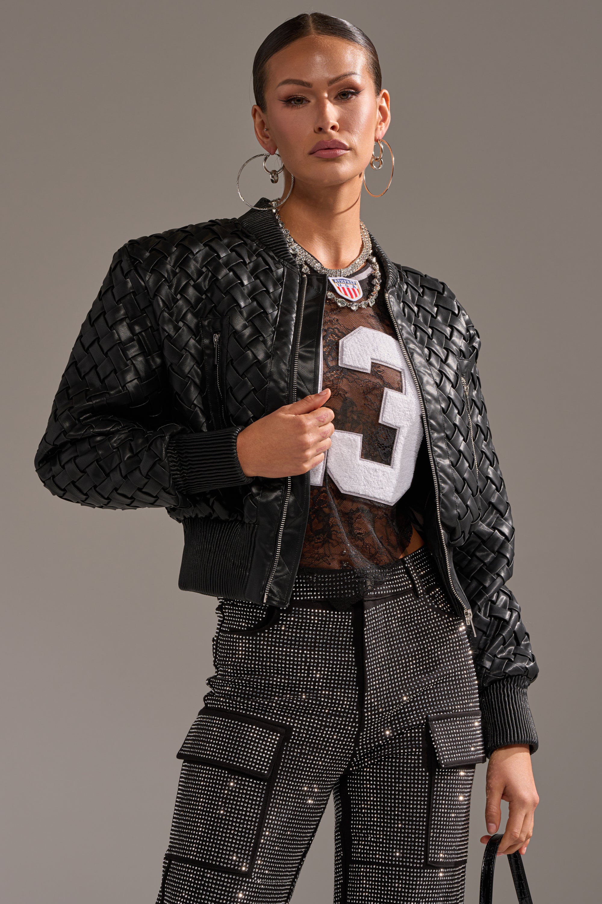 A woman poses confidently in the WICKER PARK WOVEN BOMBER, a sports jersey with the number 3, studded cargo pants, large hoop earrings, and a black handbag against a neutral background.