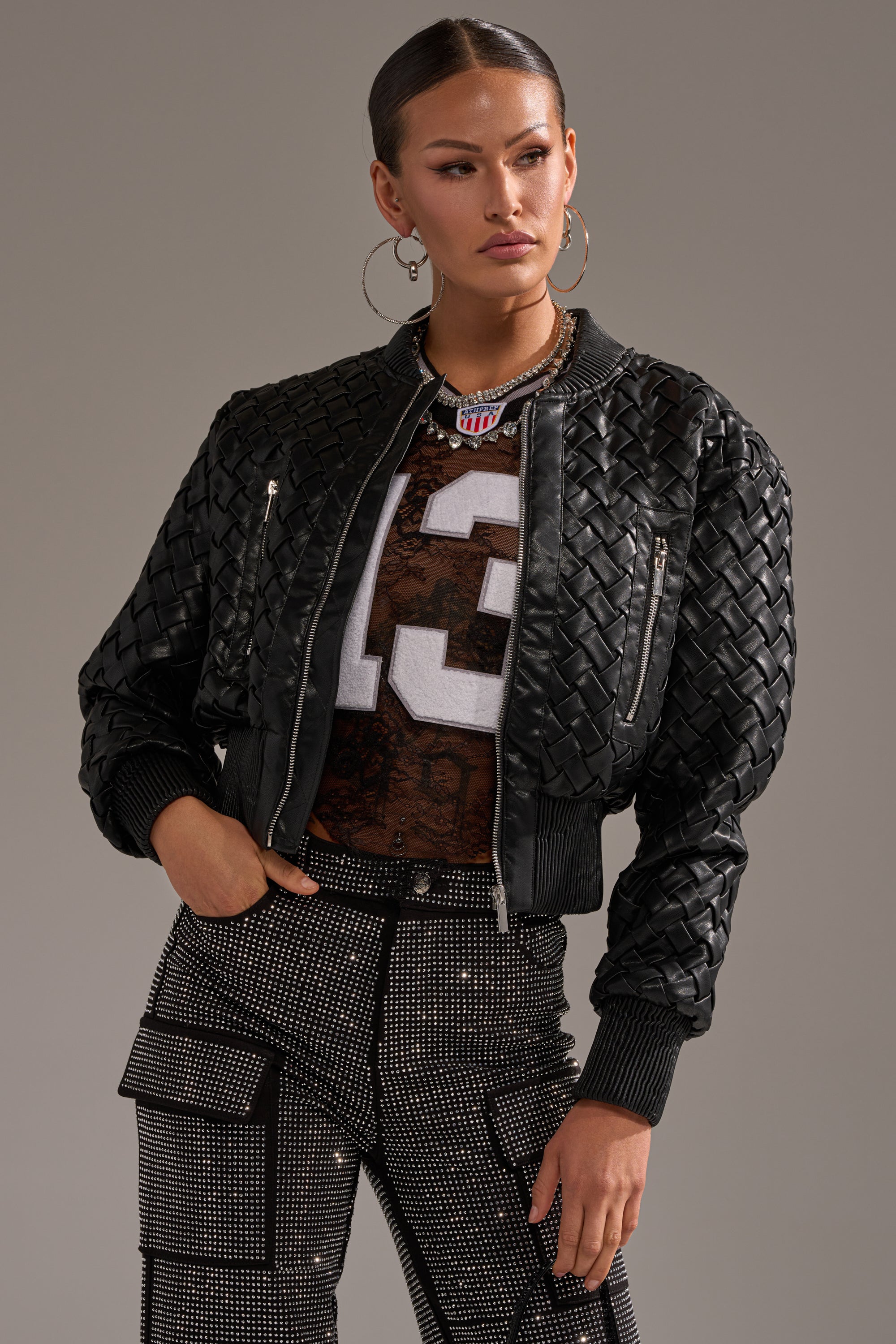 A woman models the WICKER PARK WOVEN BOMBER over a mesh football jersey and black studded pants, standing with one hand on her hip against a plain background.
