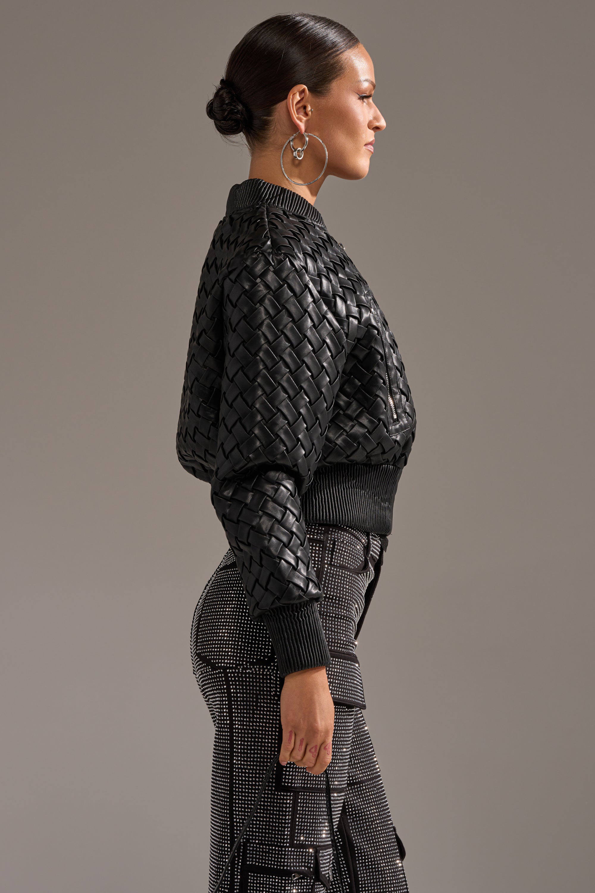 A woman with dark hair in a low bun wears the WICKER PARK WOVEN BOMBER with high-waisted, patterned pants, standing in profile against a plain gray background.